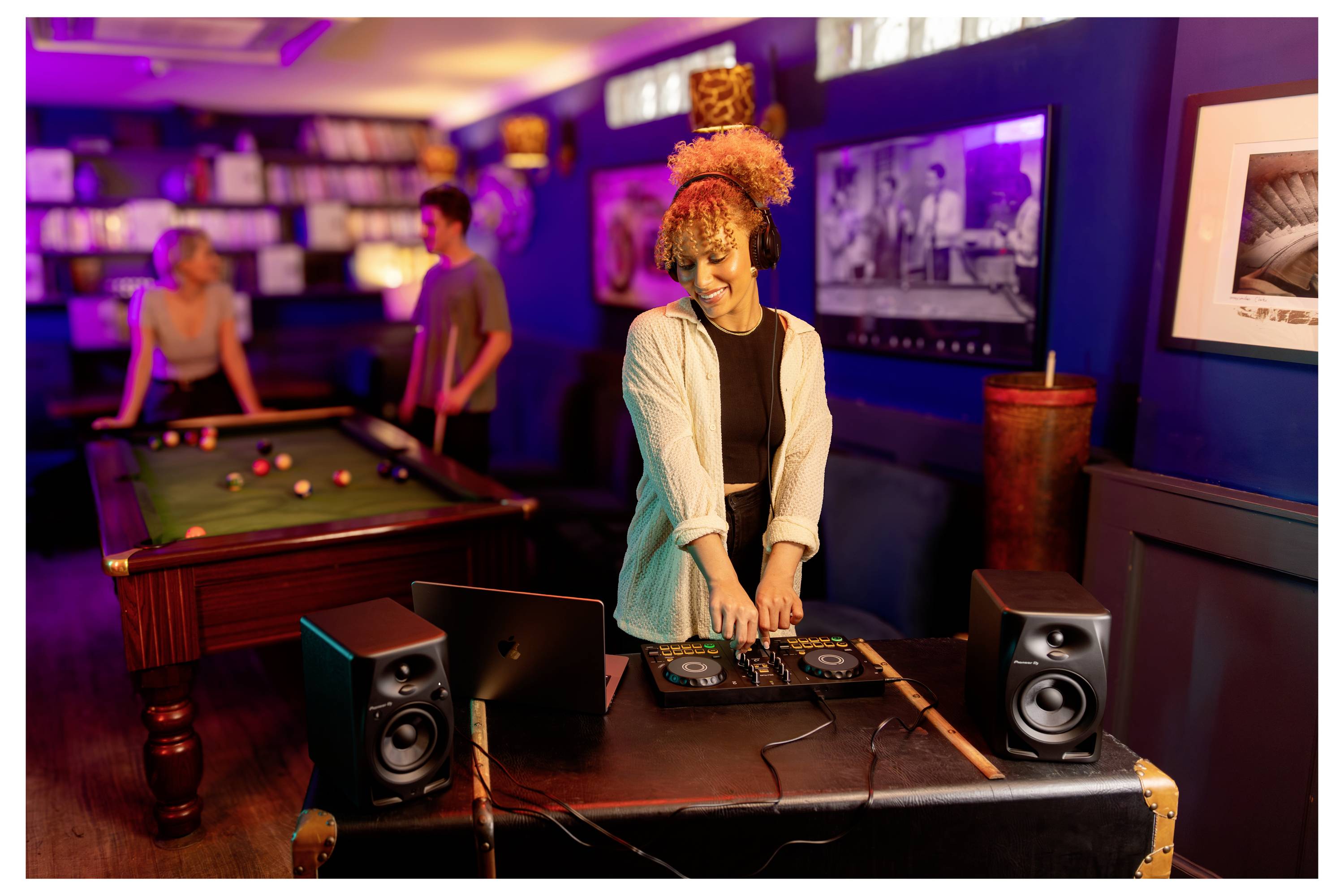 Une femme DJ mixant de la musique sur un contrôleur dans un salon, avec des gens jouant au billard en arrière-plan.