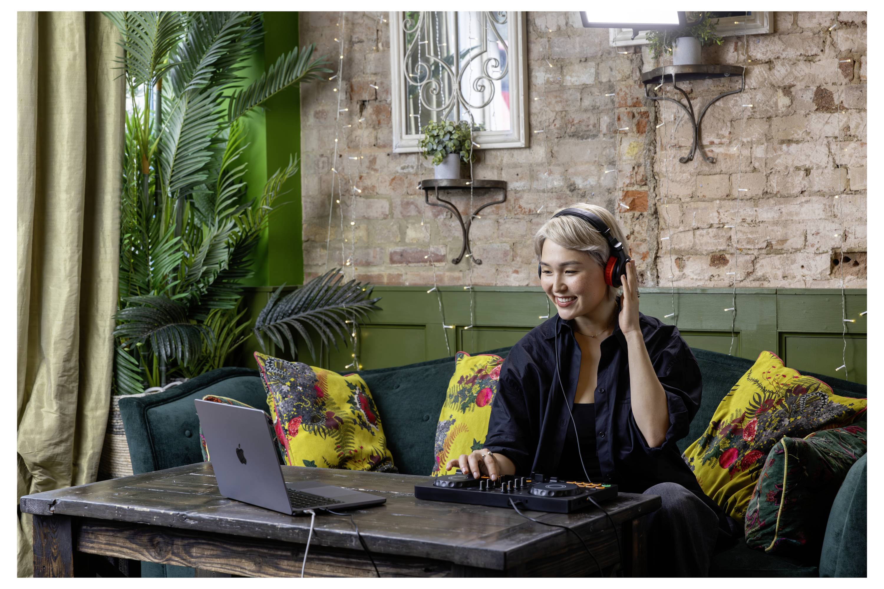 Une femme portant des écouteurs mixe l'audio sur un ordinateur portable et une console dans un lounge douillet avec un canapé et un mur en brique en arrière-plan.