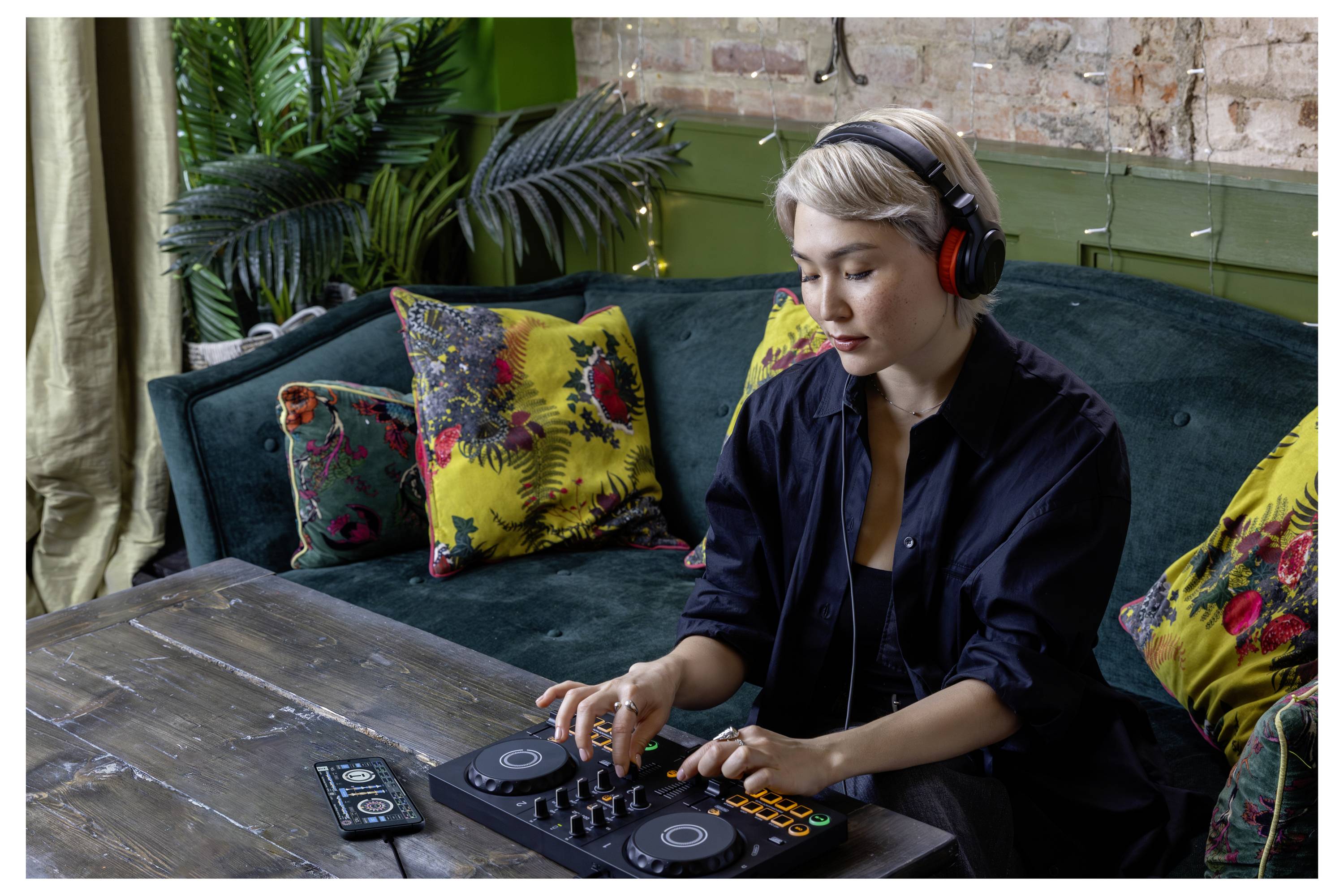 Une femme portant des écouteurs utilise une console DJ et un téléphone sur un canapé dans un salon douillet.