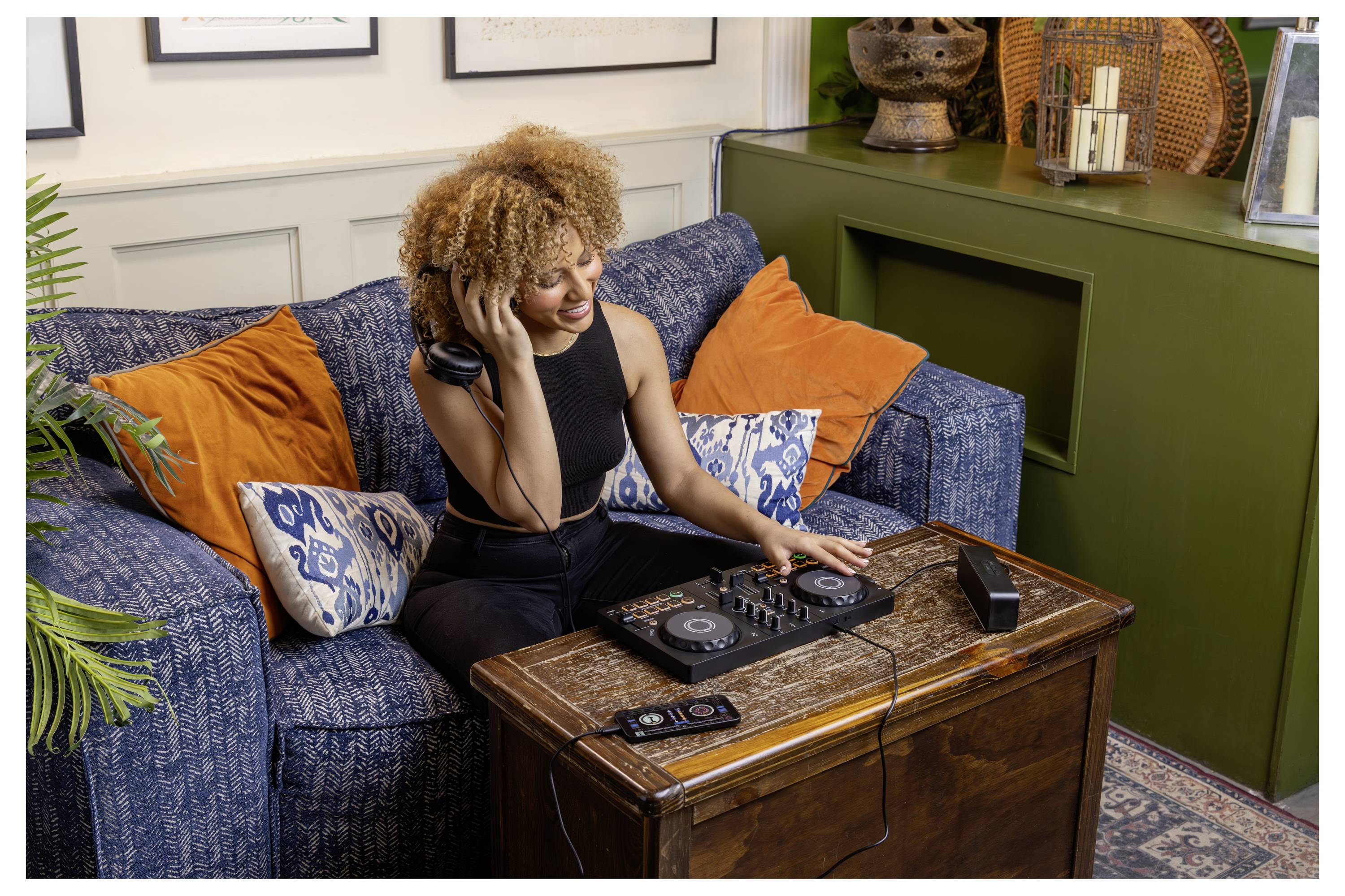 Femme portant des casques sourit en faisant du DJing sur une table basse dans un salon, assise sur un canapé bleu avec des coussins orange.