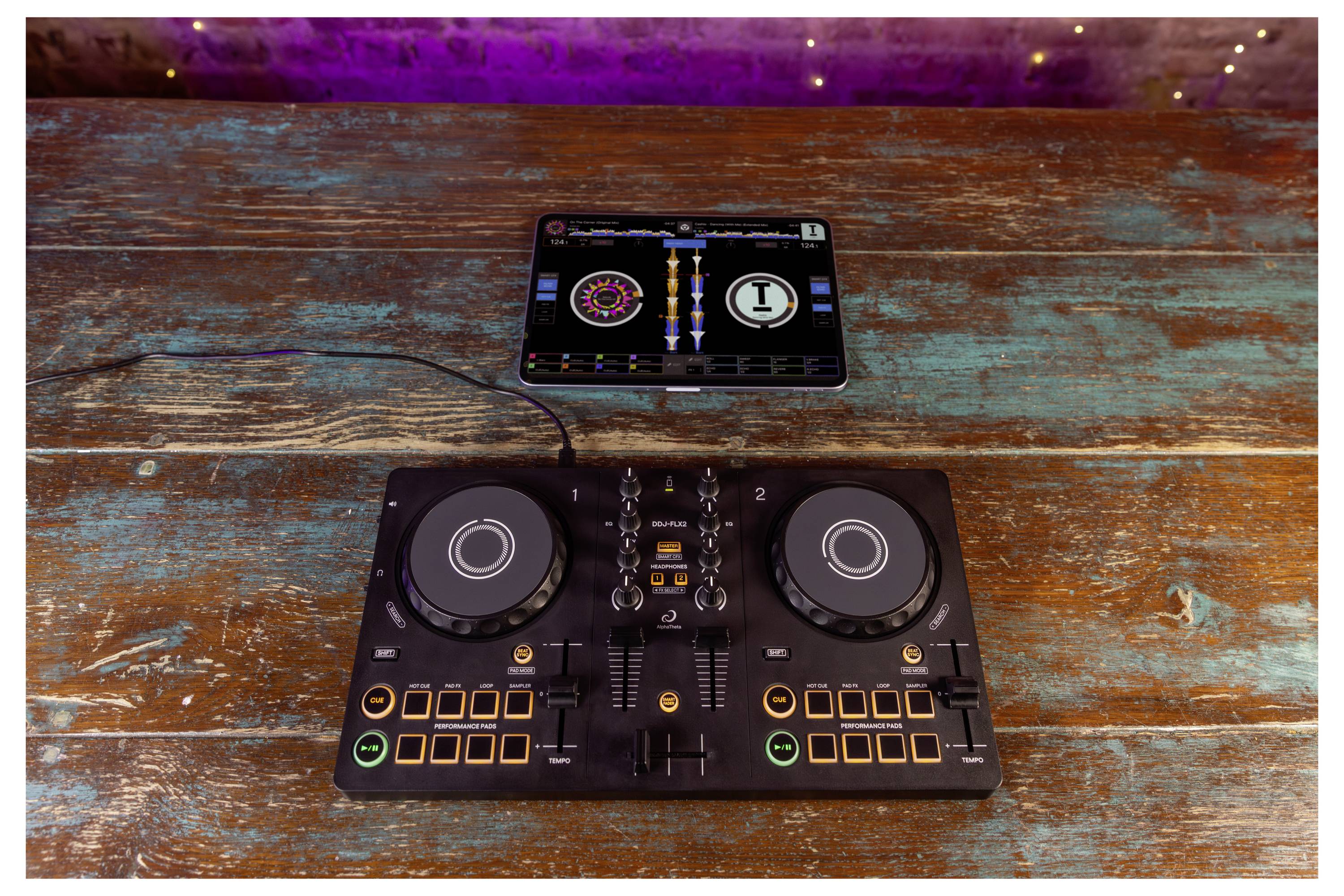 Contrôleur DJ compact sur une table en bois avec une tablette au-dessus affichant des platines virtuelles doubles et une interface de mixage centrale.