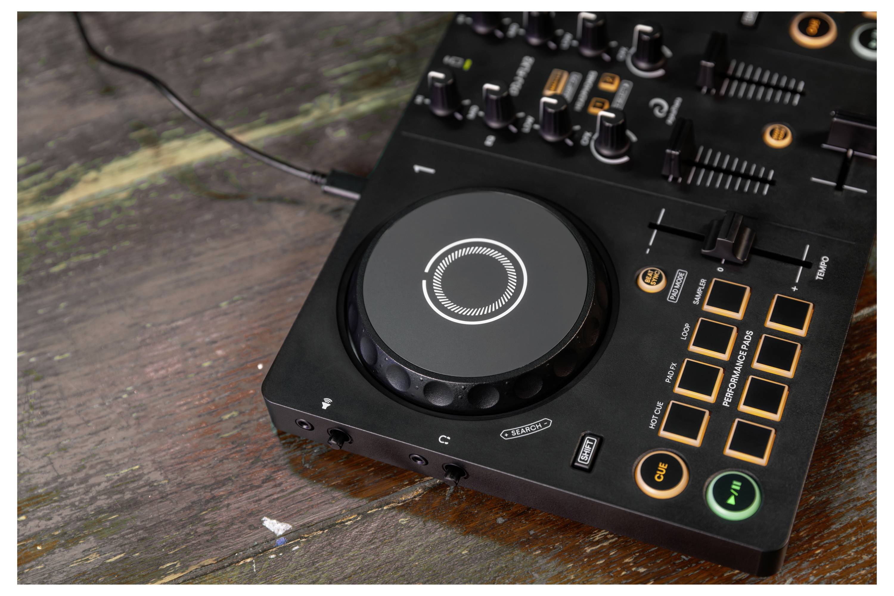 Contrôleur audio DJ noir sur une table en bois, avec une grande molette de navigation, des curseurs, des boutons rotatifs, et des pavés et boutons illuminés en orange.