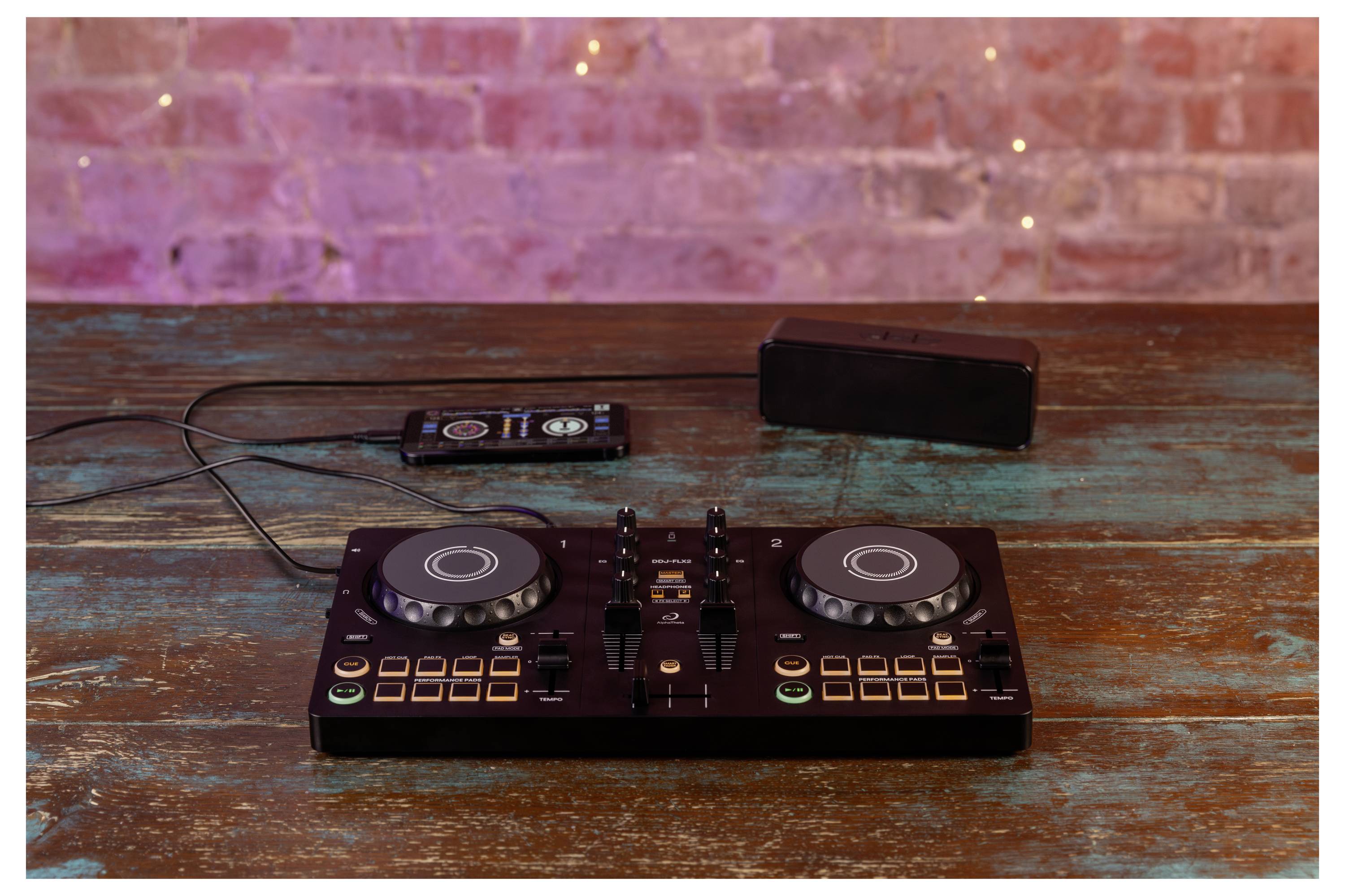 Contrôleur DJ sur une table en bois rustique, avec un téléphone, un câble et un petit haut-parleur derrière lui.