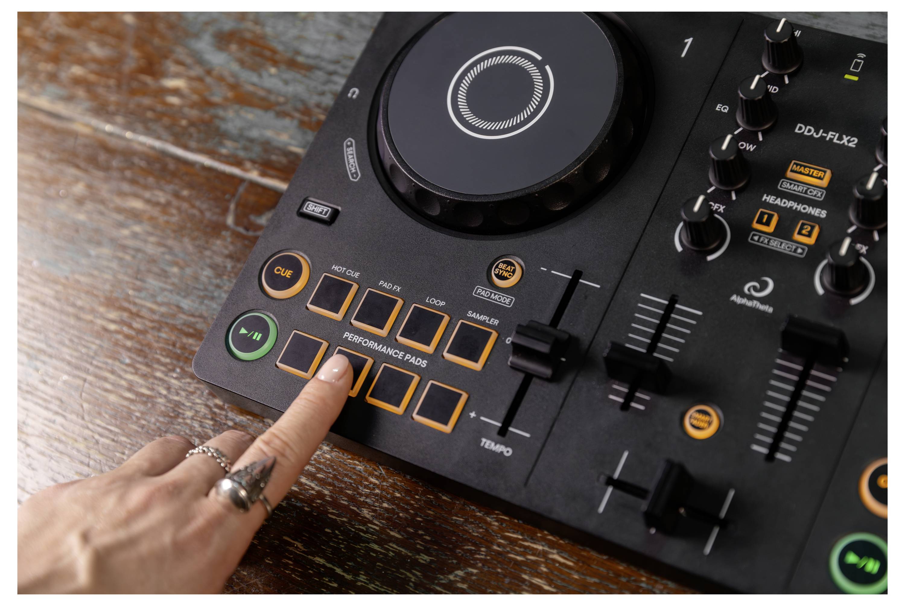 Une main appuie sur un bouton d'une manette de DJ avec un jog wheel, des pads et des curseurs sur une surface en bois.