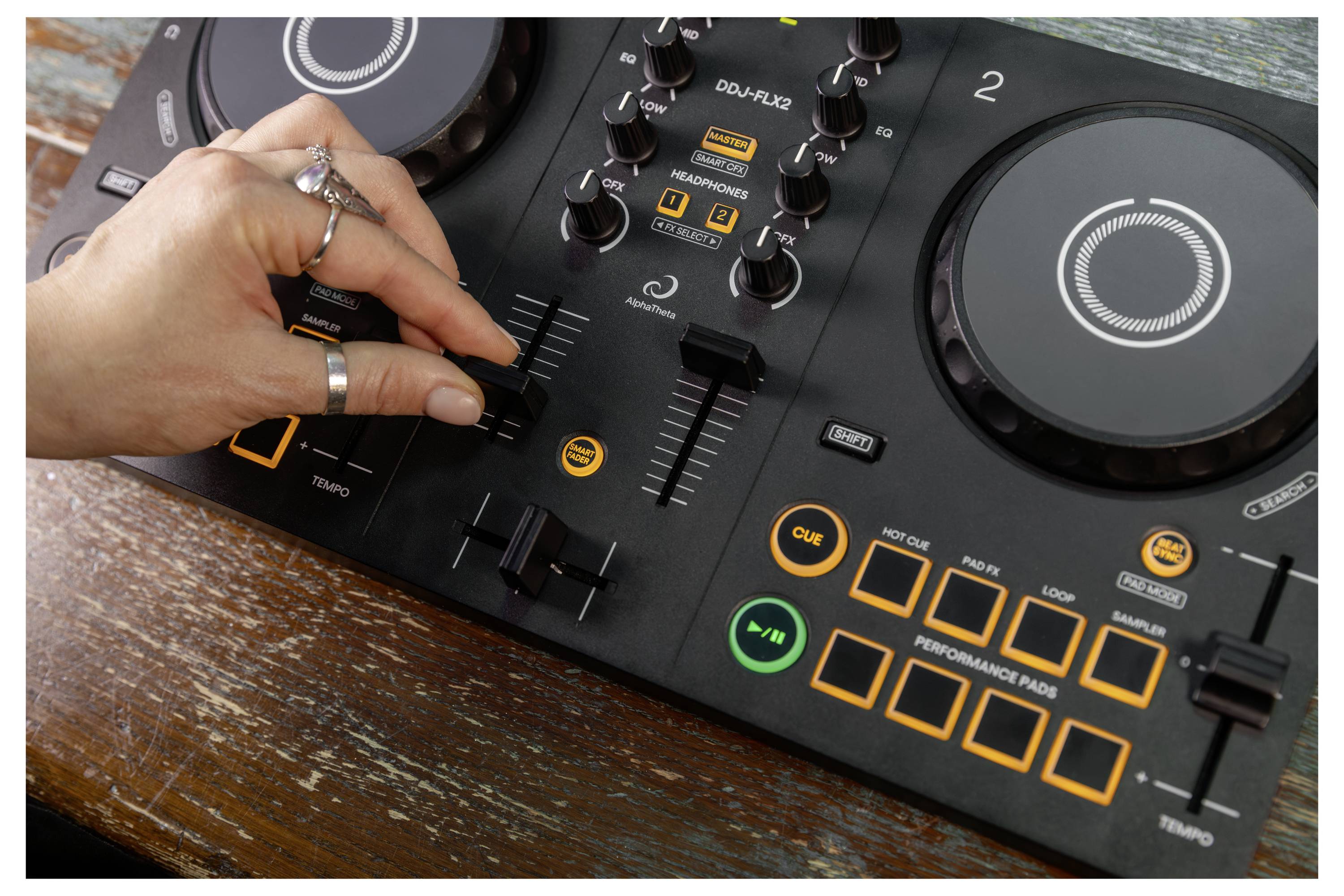 La main ajuste le crossfader sur un contrôleur DJ avec des molettes de joggage, des boutons et des pads de performance illuminés sur une table en bois.