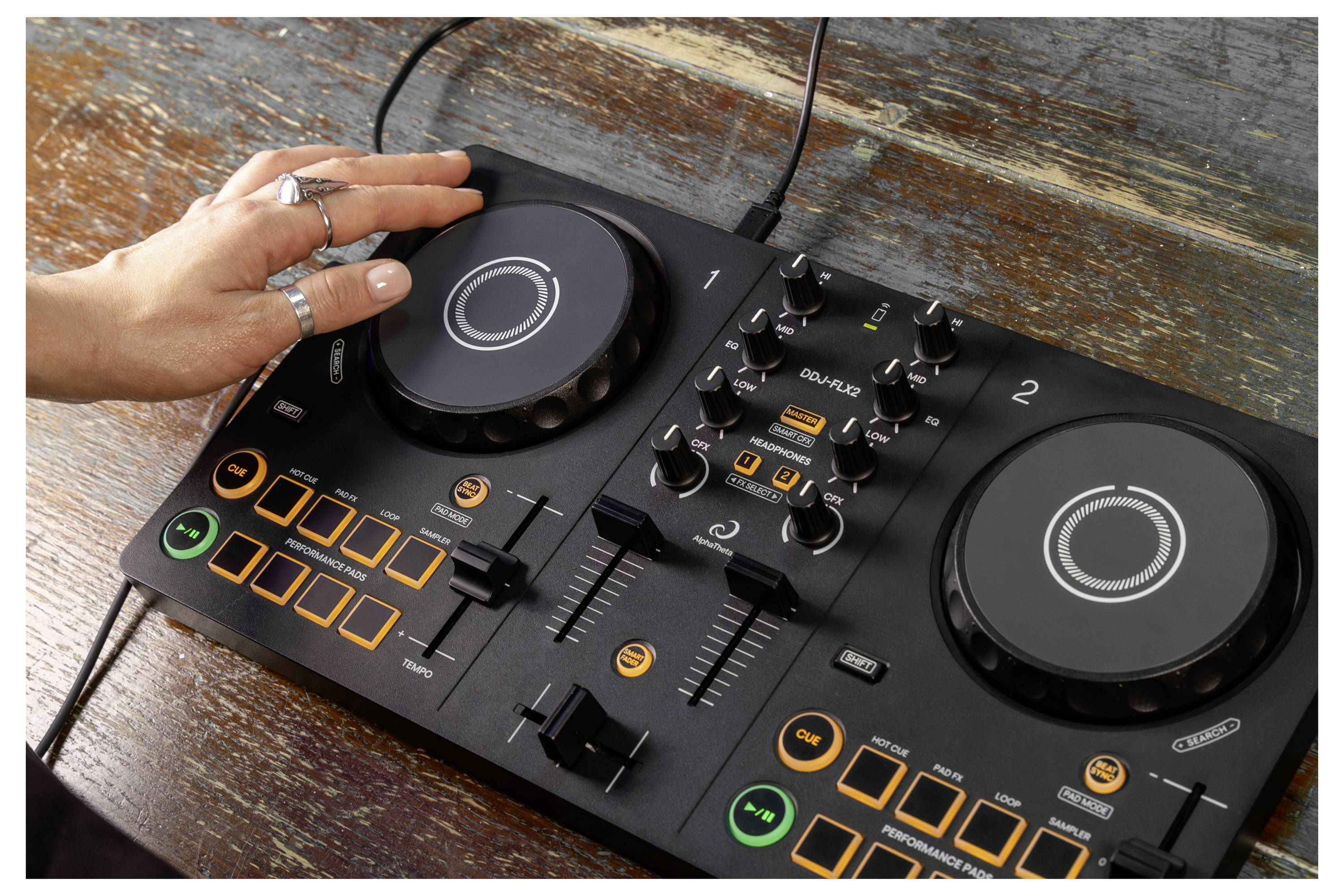 Main qui opère un contrôleur DJ compact avec deux molettes, des curseurs, des boutons de commande et des boutons de performance illuminés sur une table en bois.