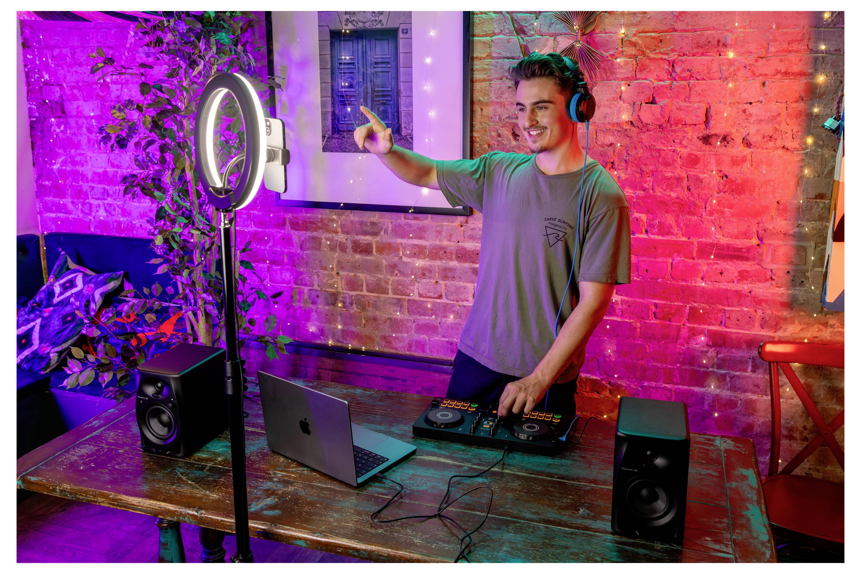 Jeune homme portant des casques audio qui fait du DJ sur un ordinateur portable et un contrôleur à côté d'une lampe annulaire dans un studio aux murs de briques colorés.