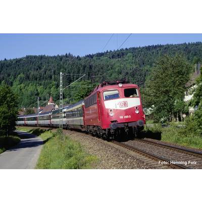 Un train de passagers DB rouge prend un virage sur des voies rurales à côté d'une route, avec des collines boisées et des maisons en arrière-plan.