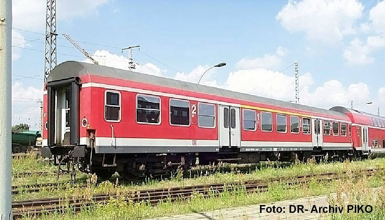 Voiture de train de passagers rouge et grise sur les rails de chemin de fer sous un ciel bleu, avec un filigrane « Foto: DR-Archiv PIKO » dans le coin.