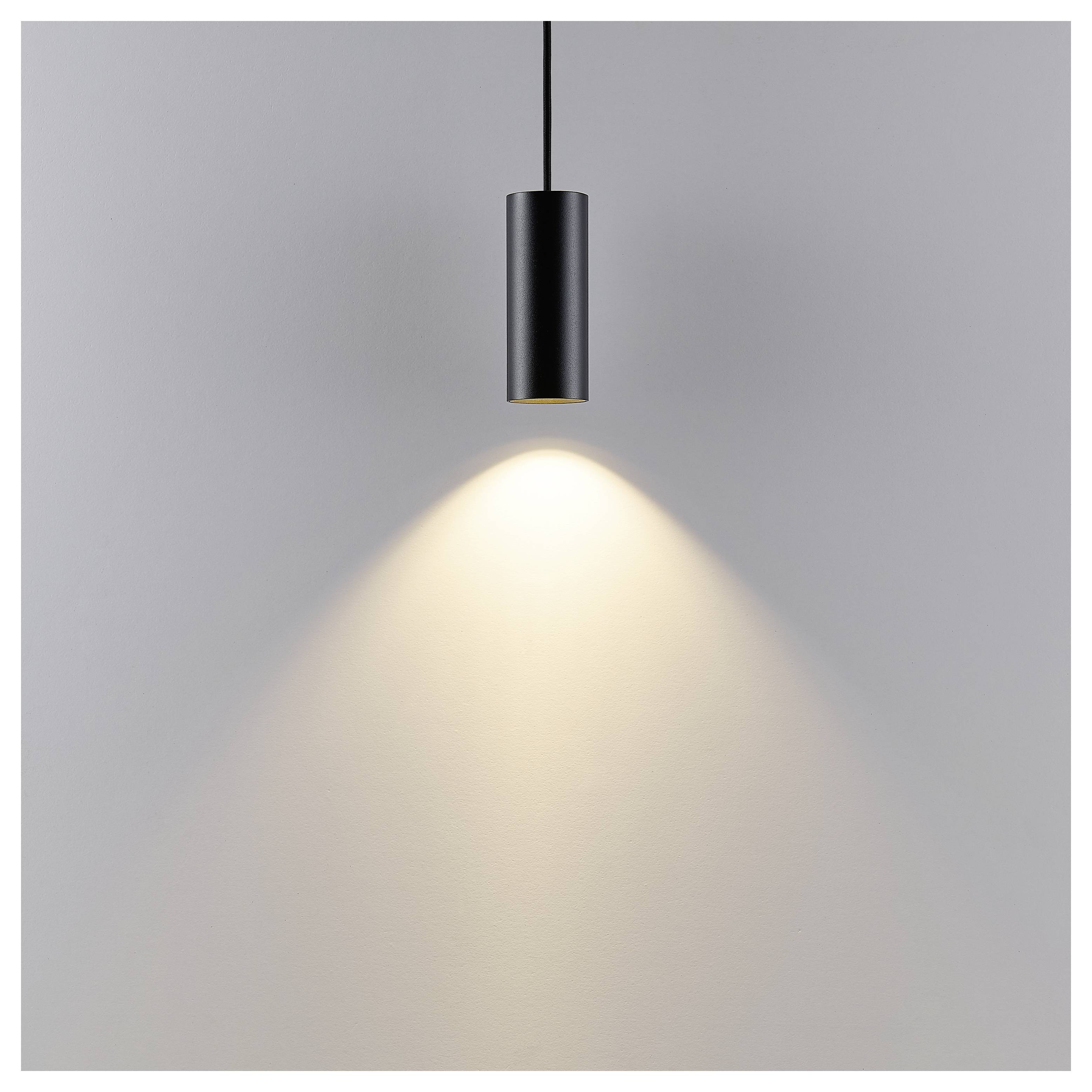 Luminaire suspendu cylindrique noir projetant un cône de lumière chaleureuse sur un mur gris uni.