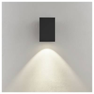 Luminaire mural noir montant en saillie projetant un cône de lumière chaud dirigé vers le bas sur un mur de couleur claire.