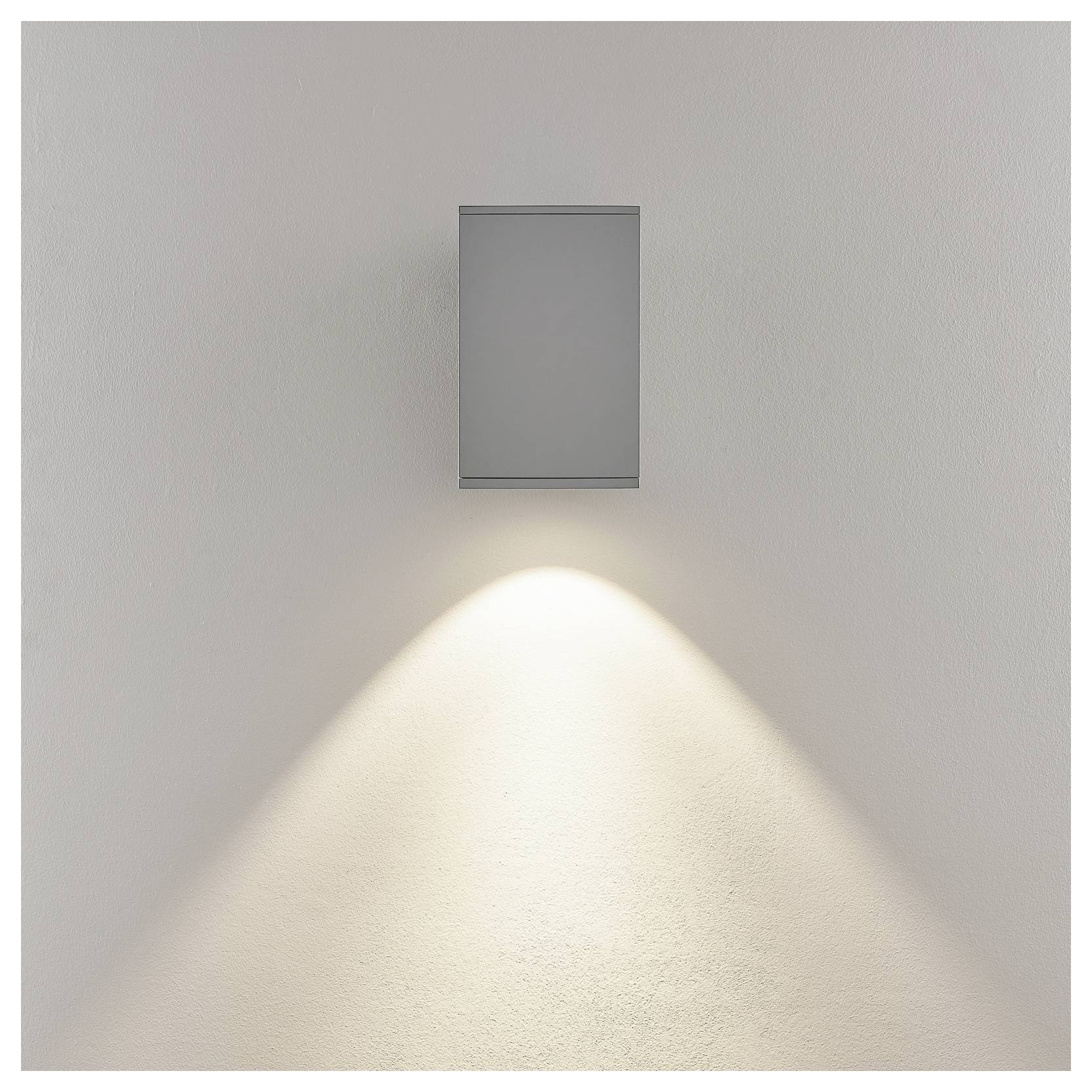 Luminaire rectangulaire mural diffusant un doux cône de lumière chaude vers le bas sur un mur blanc uni.