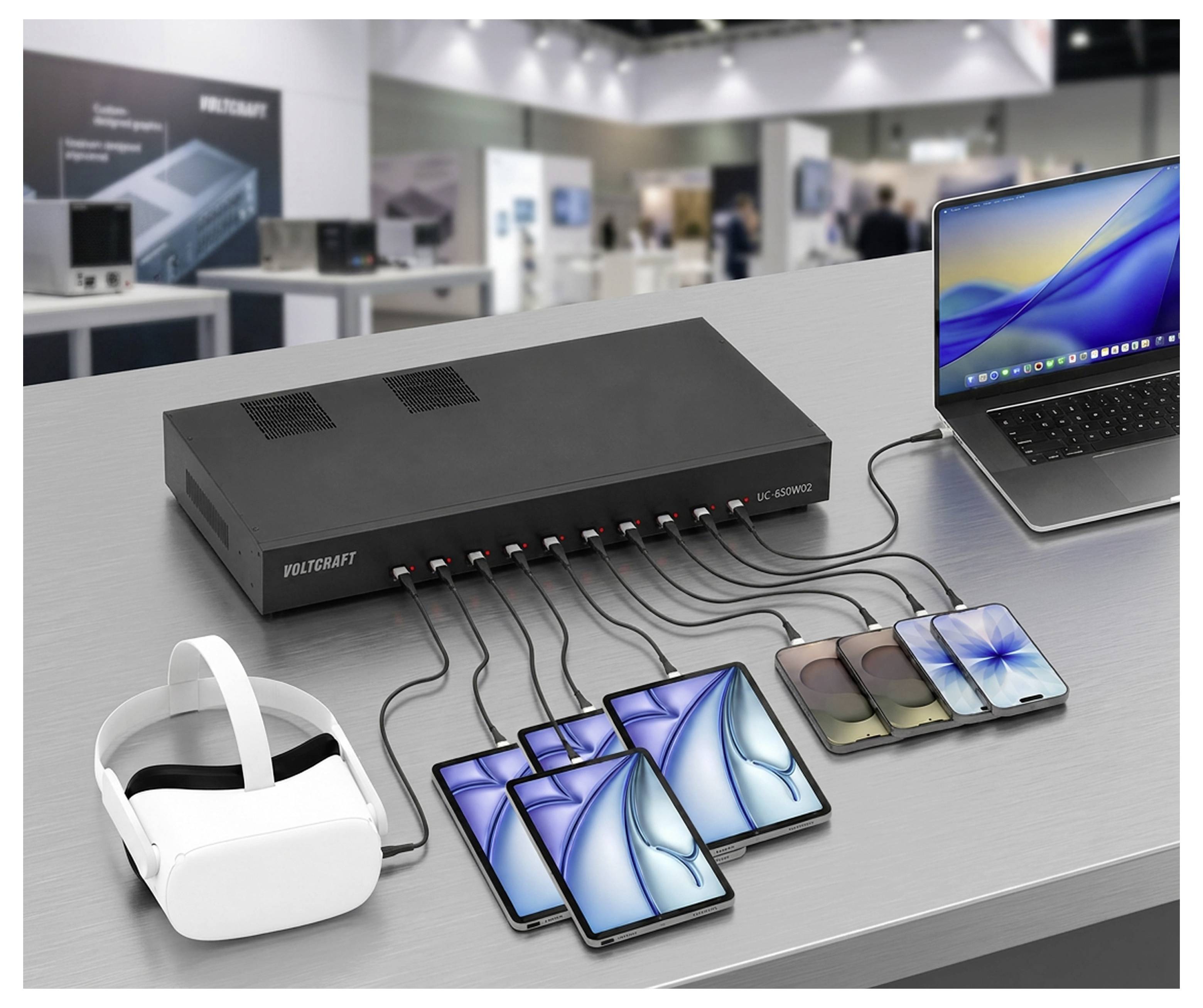Un hub de recharge USB sur une table alimenté plusieurs smartphones, un casque de réalité virtuelle et un ordinateur portable dans un affichage de salon commercial.