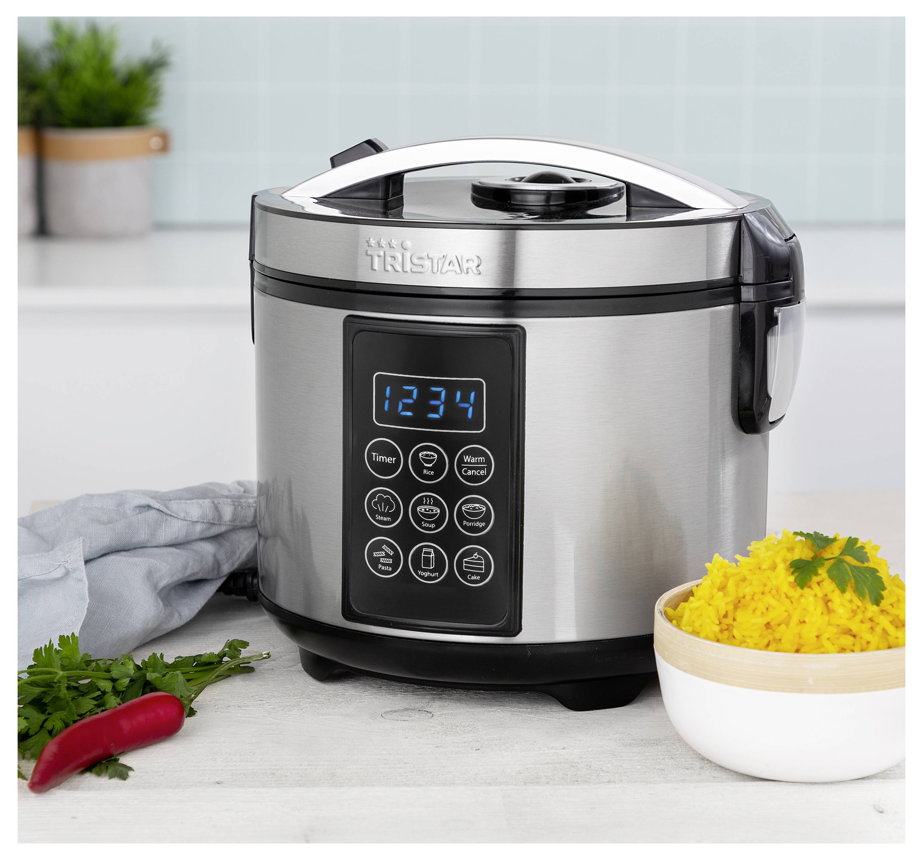 Un multicuiseur Tristar en acier inoxydable avec panneau de commande numérique est posé sur un plan de travail de cuisine à côté d'un bol de riz, d'herbes aromatiques et d'un piment rouge.