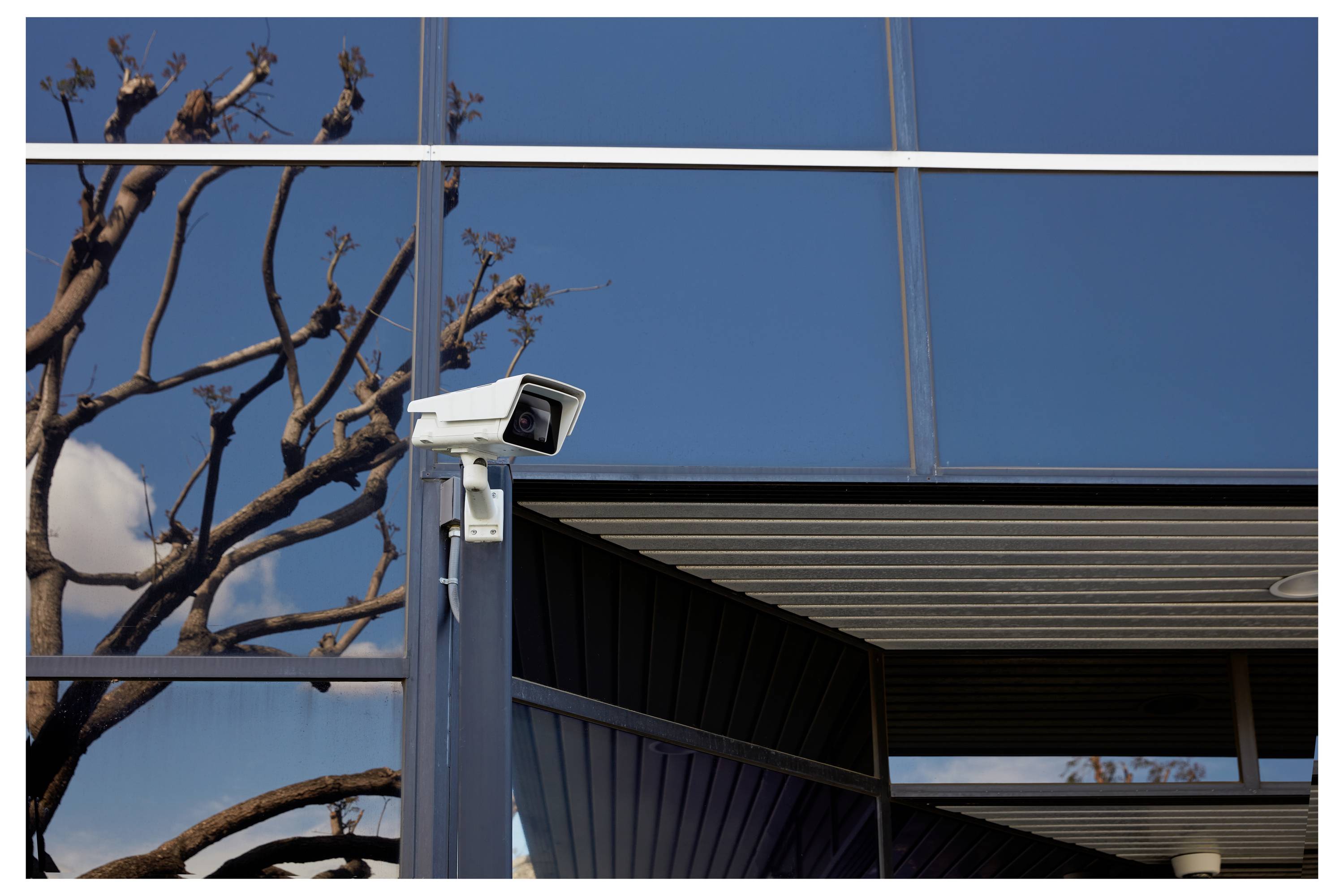 Caméra de surveillance montée sur l'extérieur d'un bâtiment bleu avec des fenêtres réfléchissantes et des branches d'arbres dénudées au premier plan.