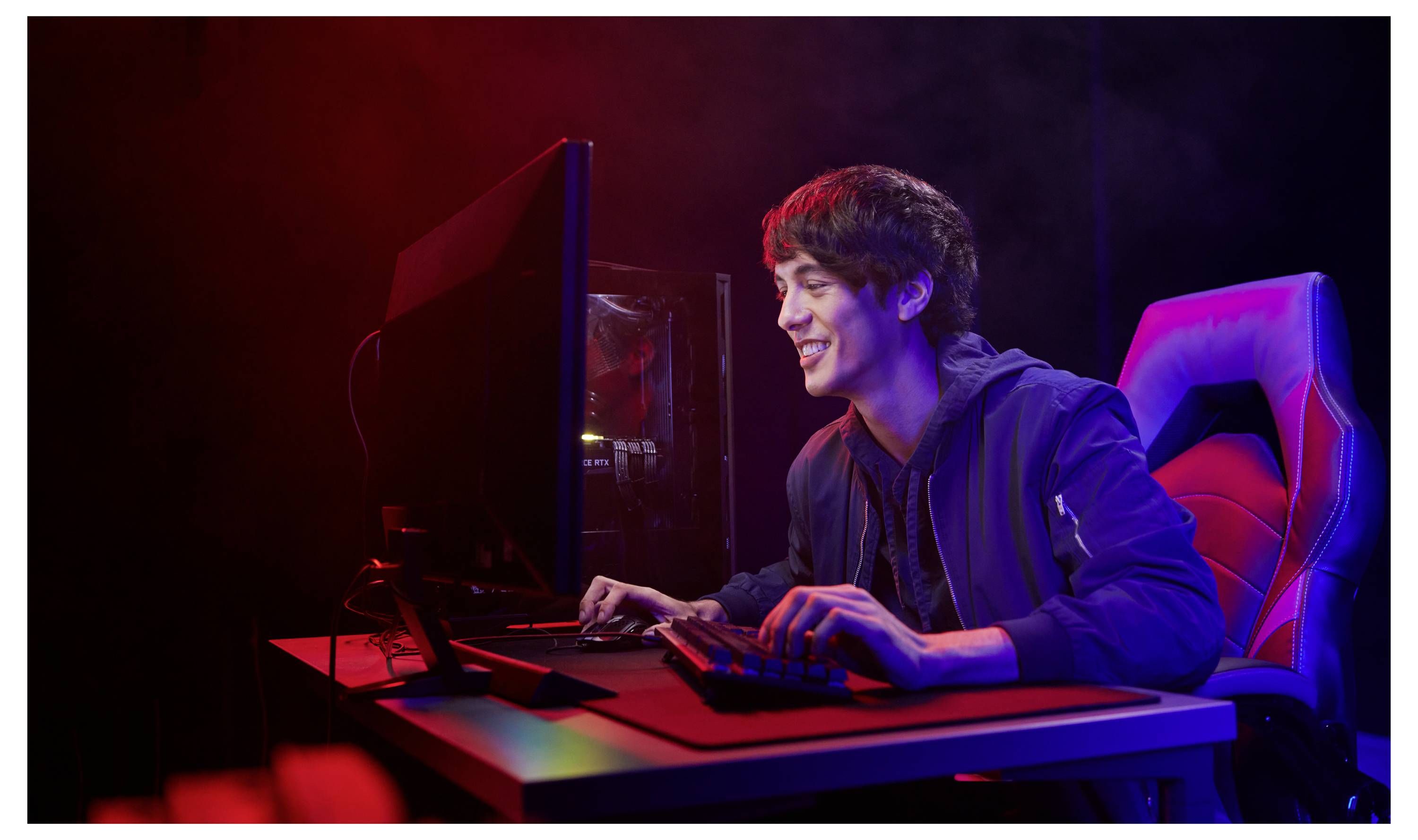 Jeune homme souriante en train de jouer à des jeux vidéo à un ordinateur de bureau dans un environnement éclairé aux néons, assis sur une chaise de jeu rouge et bleue.