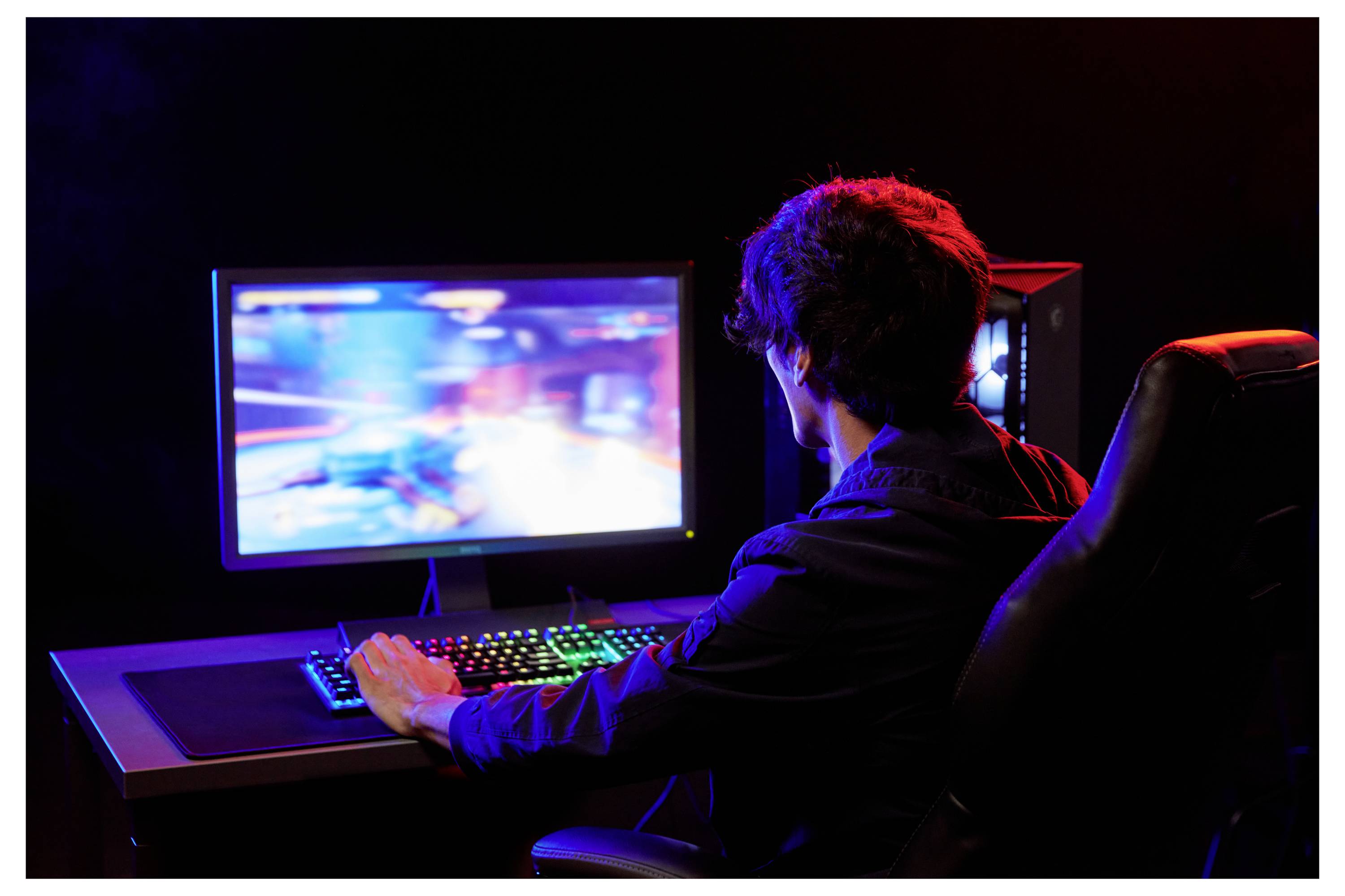 Personne jouant sur un ordinateur de bureau dans une pièce sombre, vue de dos, avec un écran et un clavier illuminés et colorés.