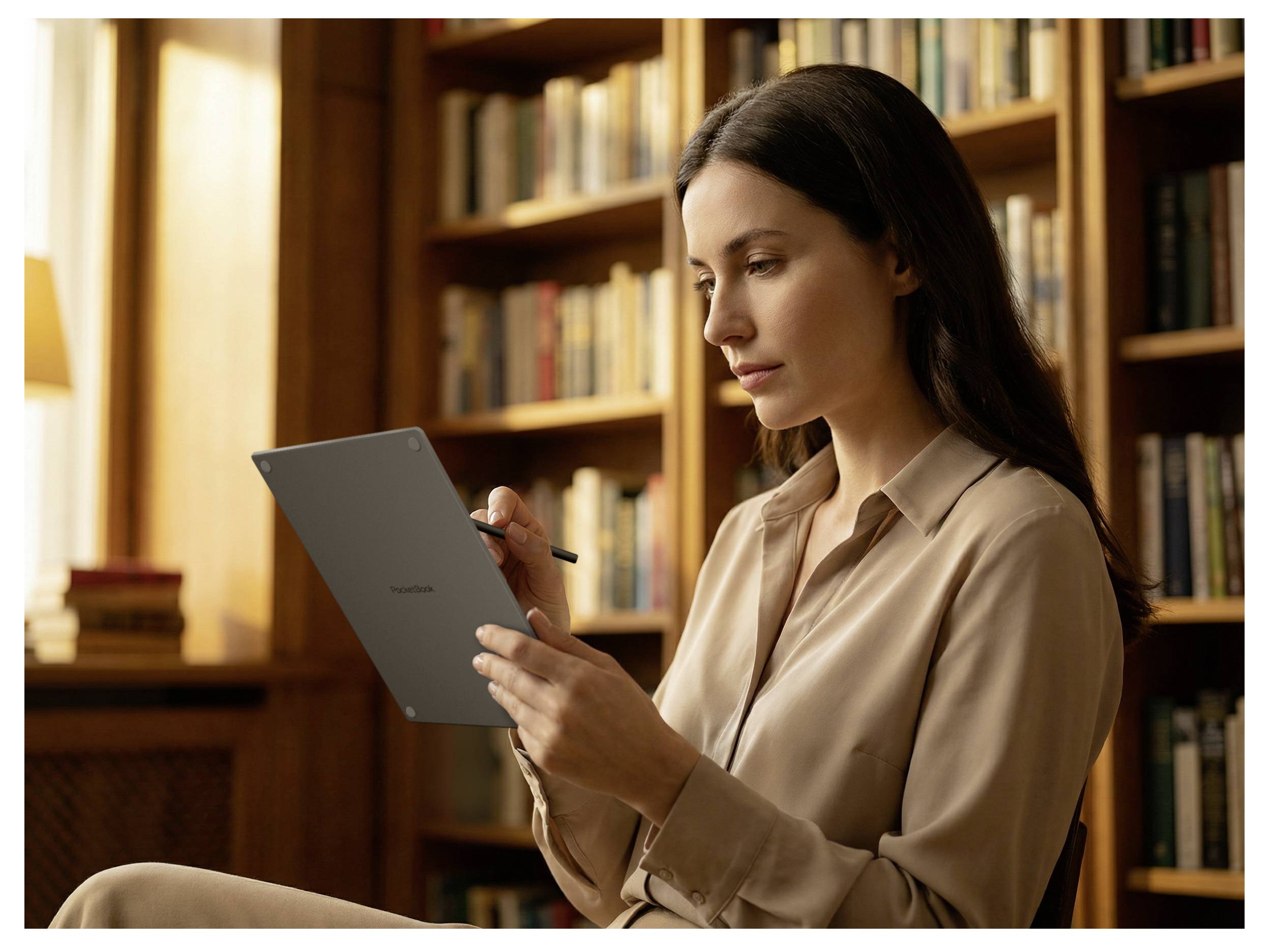 Femme assise dans une bibliothèque, écrivant sur une tablette avec un stylet tout en lisant, avec des rayons de livres en arrière-plan.