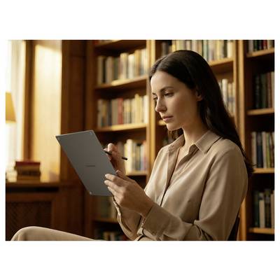 Femme assise dans une bibliothèque, écrivant sur une tablette avec un stylet tout en lisant, avec des rayons de livres en arrière-plan.