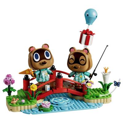 Deux personnages ours sur un pont rouge enjambant un ruisseau bleu, l'un pêchant tandis qu'un cadeau descend en ballon ; scène de style LEGO avec des fleurs et des plantes.