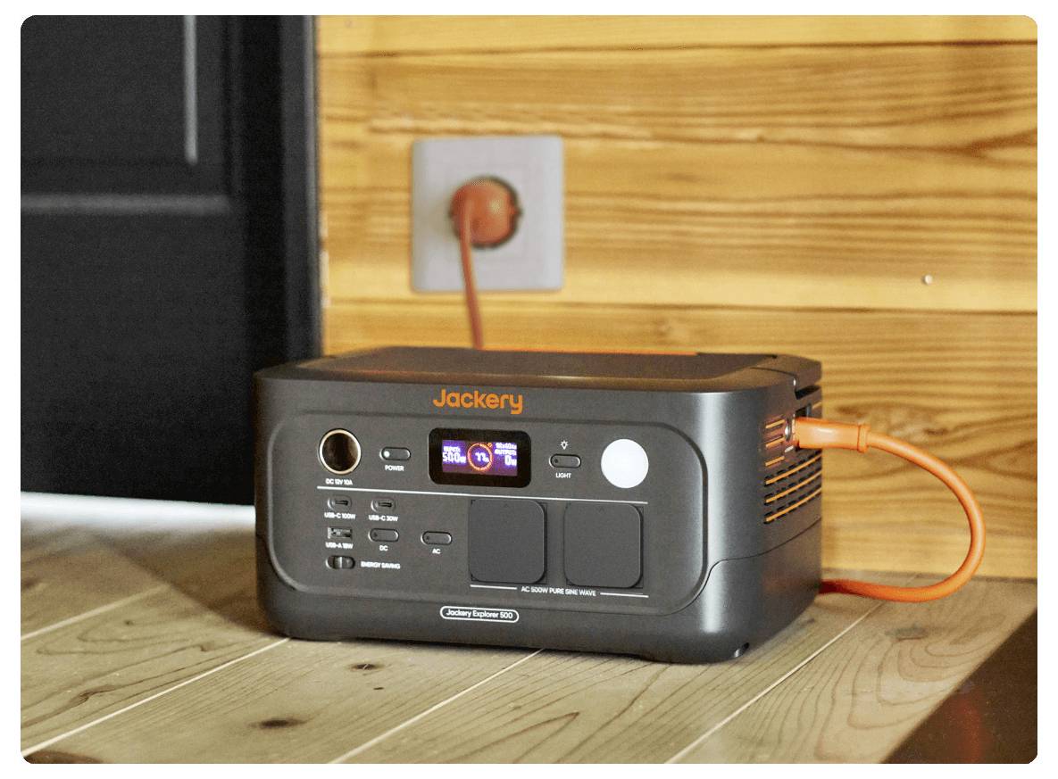 Station d'énergie portable Jackery branchée sur une prise murale, posée sur un plancher en bois contre un mur recouvert de boiseries.