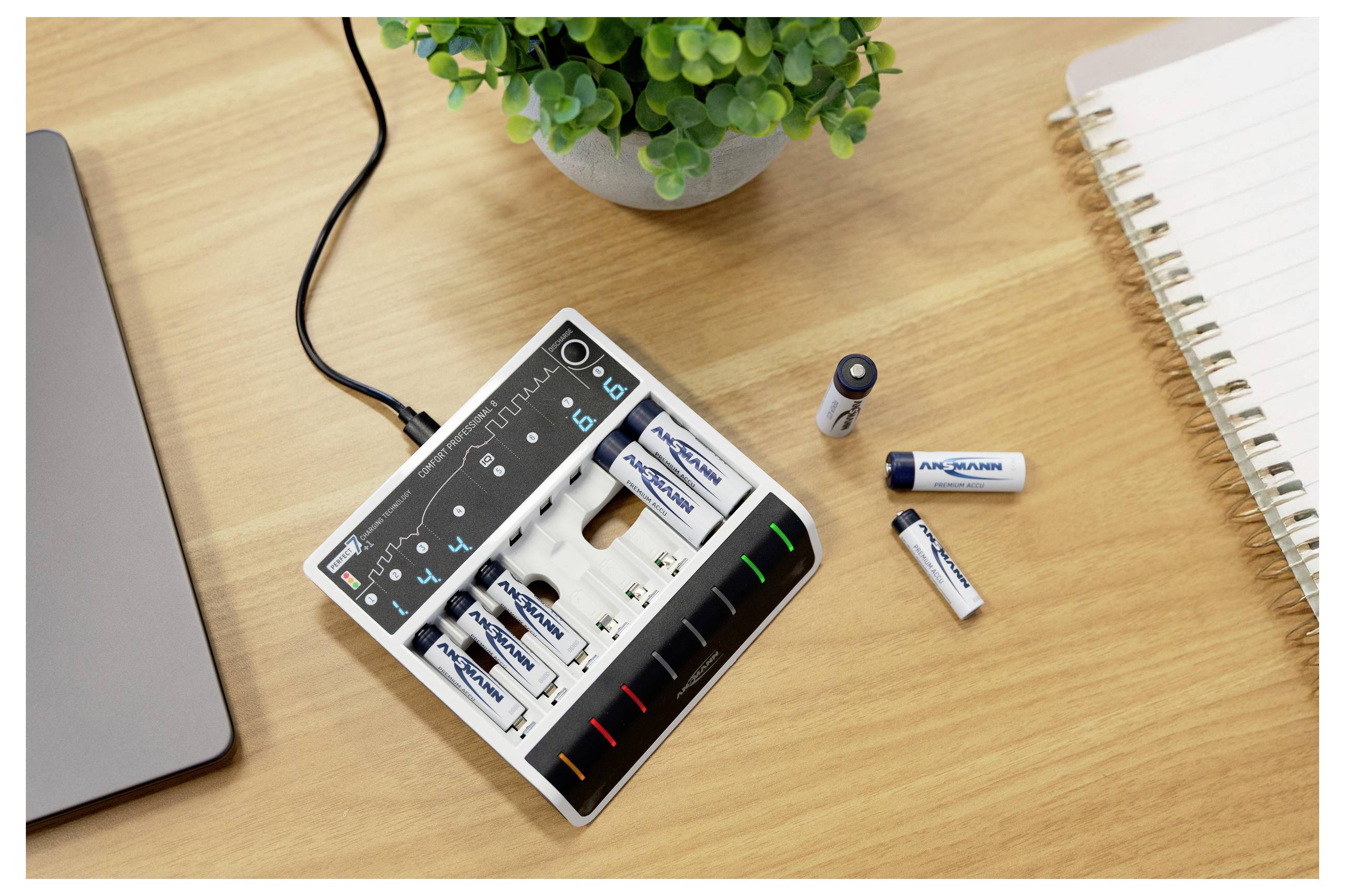 Chargeur de piles AA et AAA avec plusieurs piles sur un bureau en bois à côté d'une plante, d'un carnet et du bord d'un ordinateur portable.