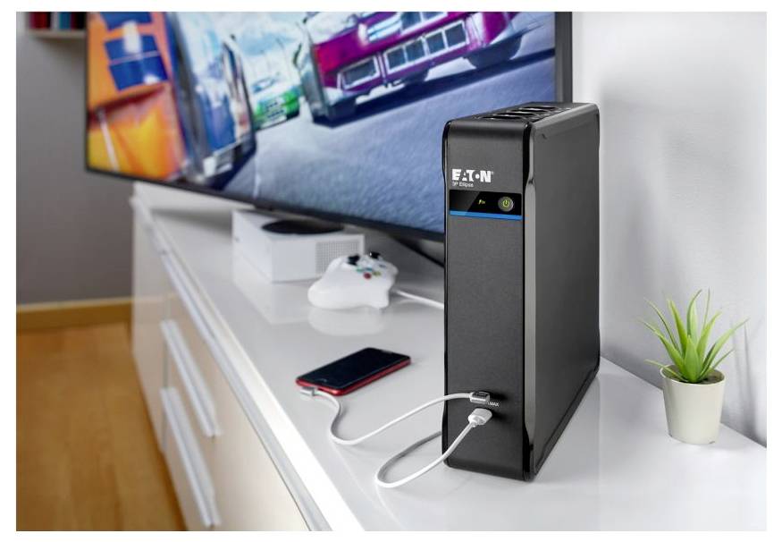 Unité de secours batterie Eaton à côté d'un téléviseur et d'une manette de jeu sur une console blanche, alimentant un smartphone avec un câble de charge.