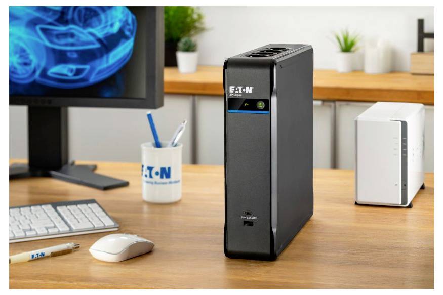 Unité de sauvegarde batterie Eaton noire sur un bureau à côté d'un écran d'ordinateur, d'un clavier, d'une souris et d'un appareil réseau blanc dans un environnement de bureau.