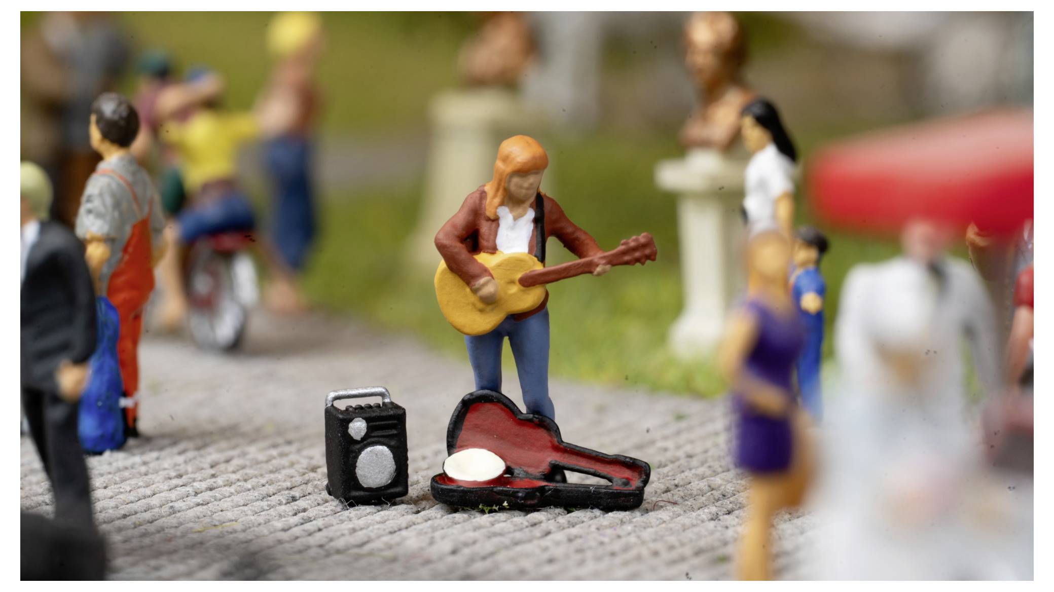 Figurine miniature de musicien de rue jouant de la guitare à côté d'un étui à instrument ouvert et d'un petit amplificateur, avec des piétons flous en avant-plan et en arrière-plan.