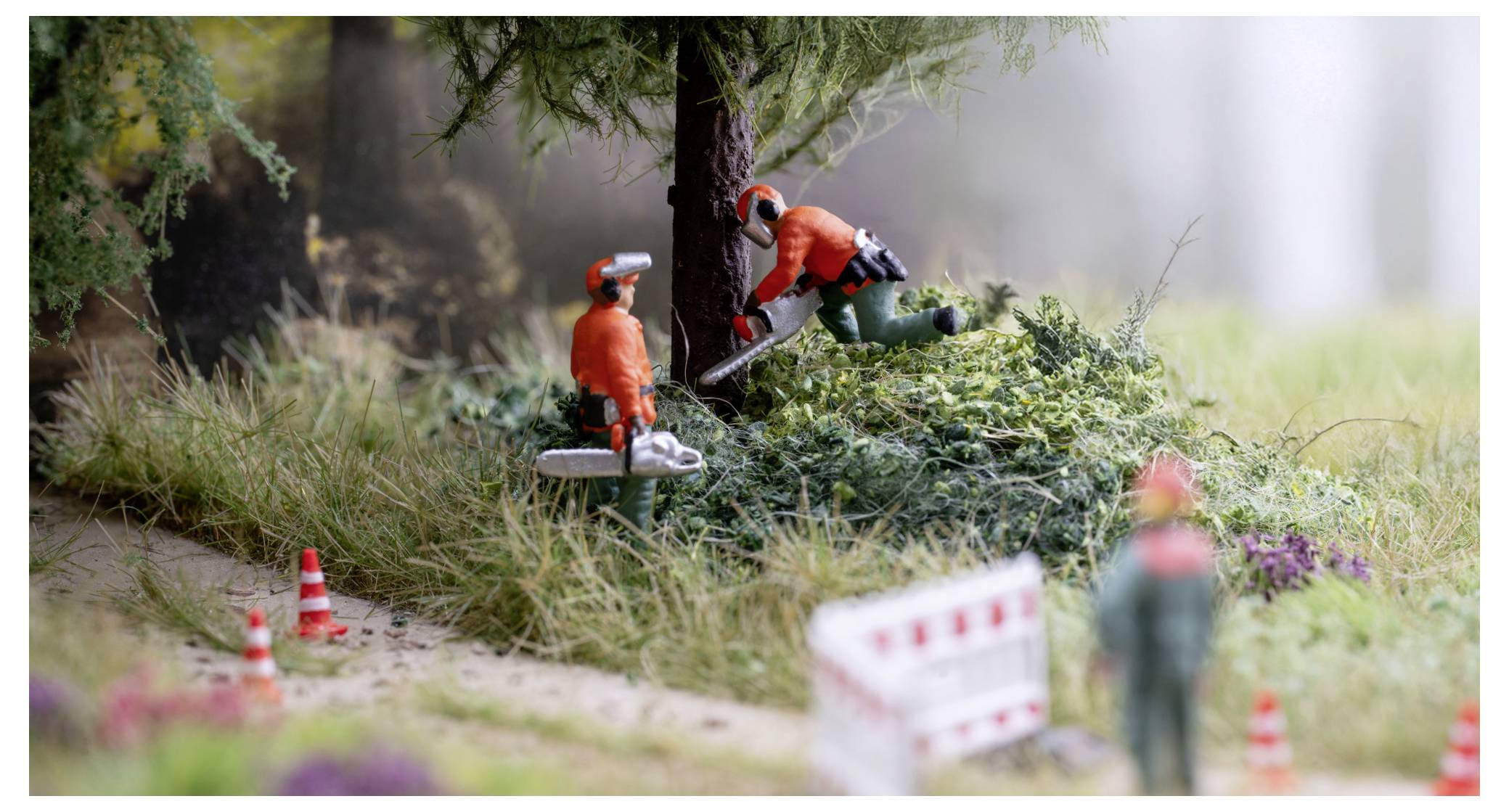 Des travailleurs miniatures en équipement de sécurité orange taillent un arbre avec des tronçonneuses à côté d'un sentier, avec des cônes de circulation et des barrières marquant la zone de travail.