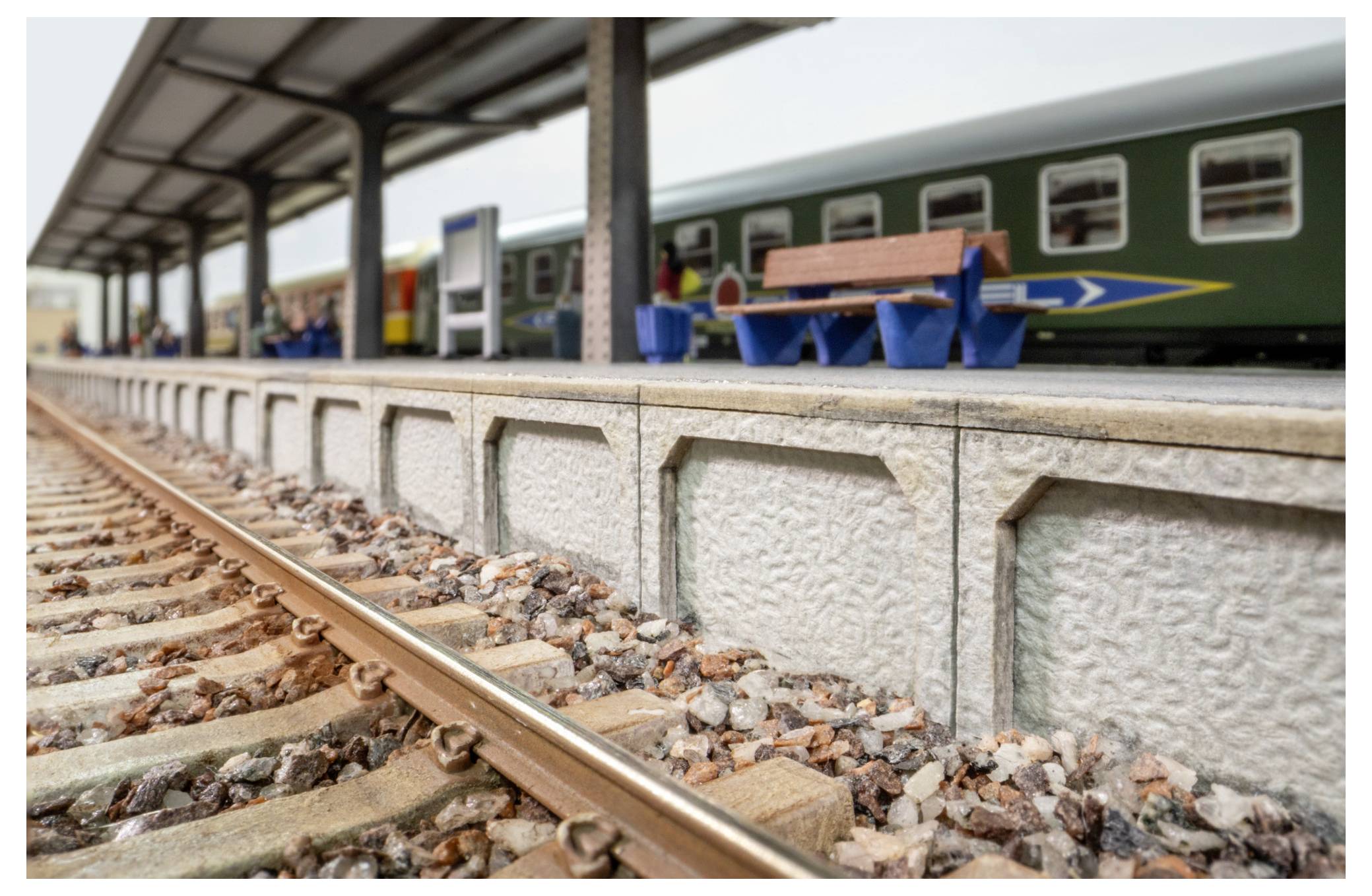 Quai de train miniature à côté d'une voie ferrée, avec une voiture de passagers verte et un banc sous un auvent de gare couvert.