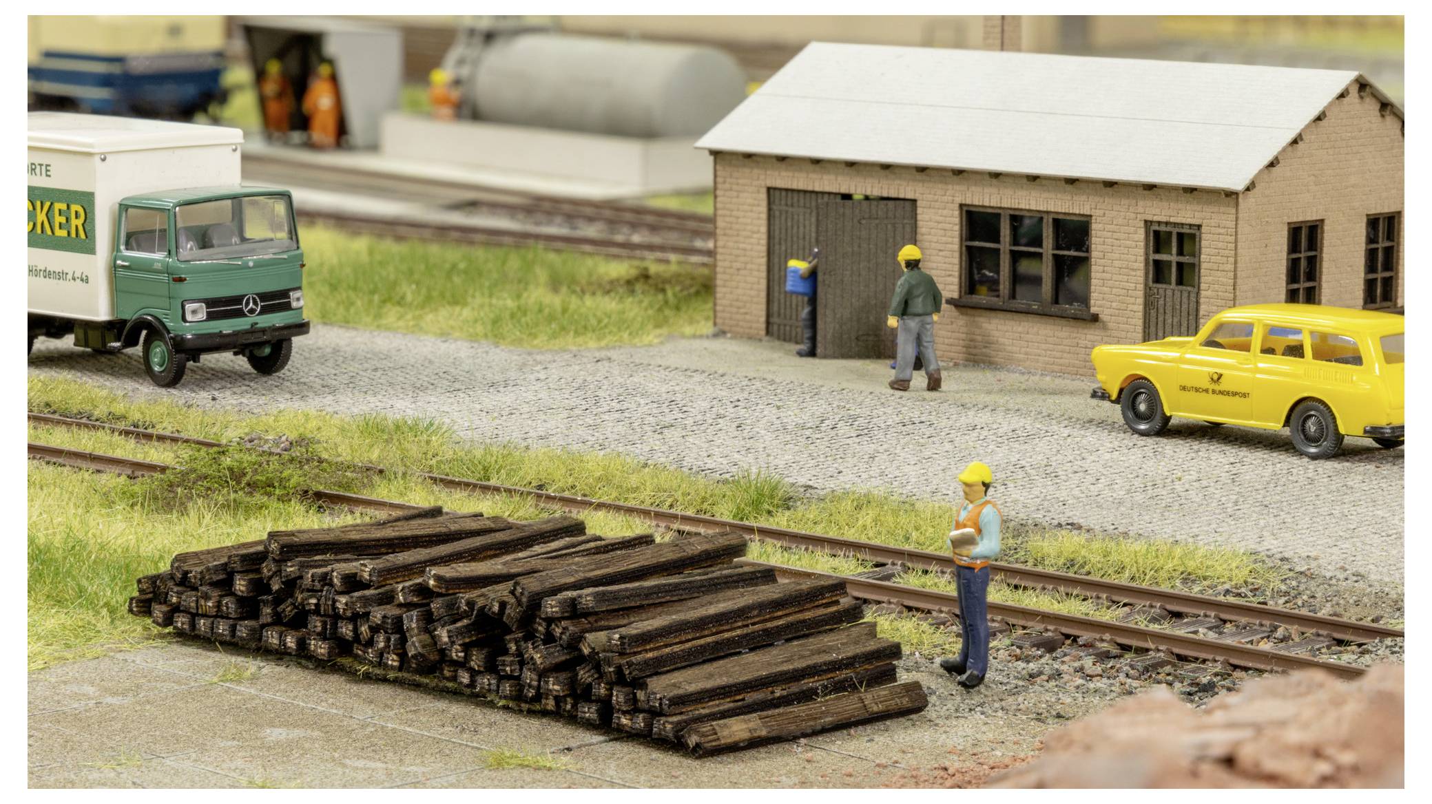 Scène de train miniature avec des ouvriers déchargeant un wagon à côté d'un dépôt en briques, une voiture jaune stationnée à proximité, et du bois empilé au premier plan.