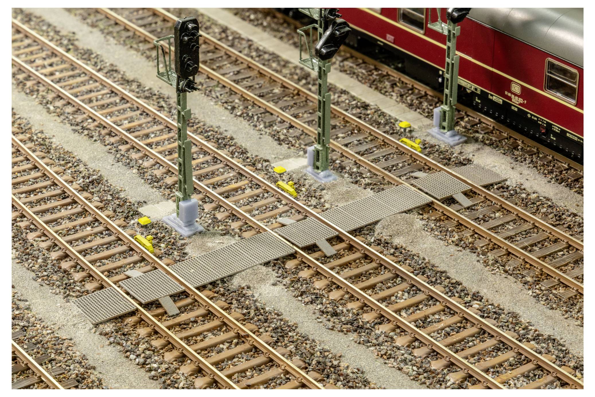 Les voies ferrées d'un chemin de fer miniature se croisent à un carrefour de niveau avec des mâts de signalisation, tandis qu'un train de passagers rouge est partiellement visible en haut à droite.