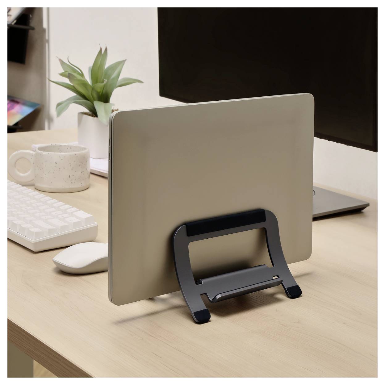 Un ordinateur portable en argent fermé repose verticalement sur un support noir sur un bureau en bois à côté d'un clavier, d'une souris, d'une tasse et d'un moniteur.