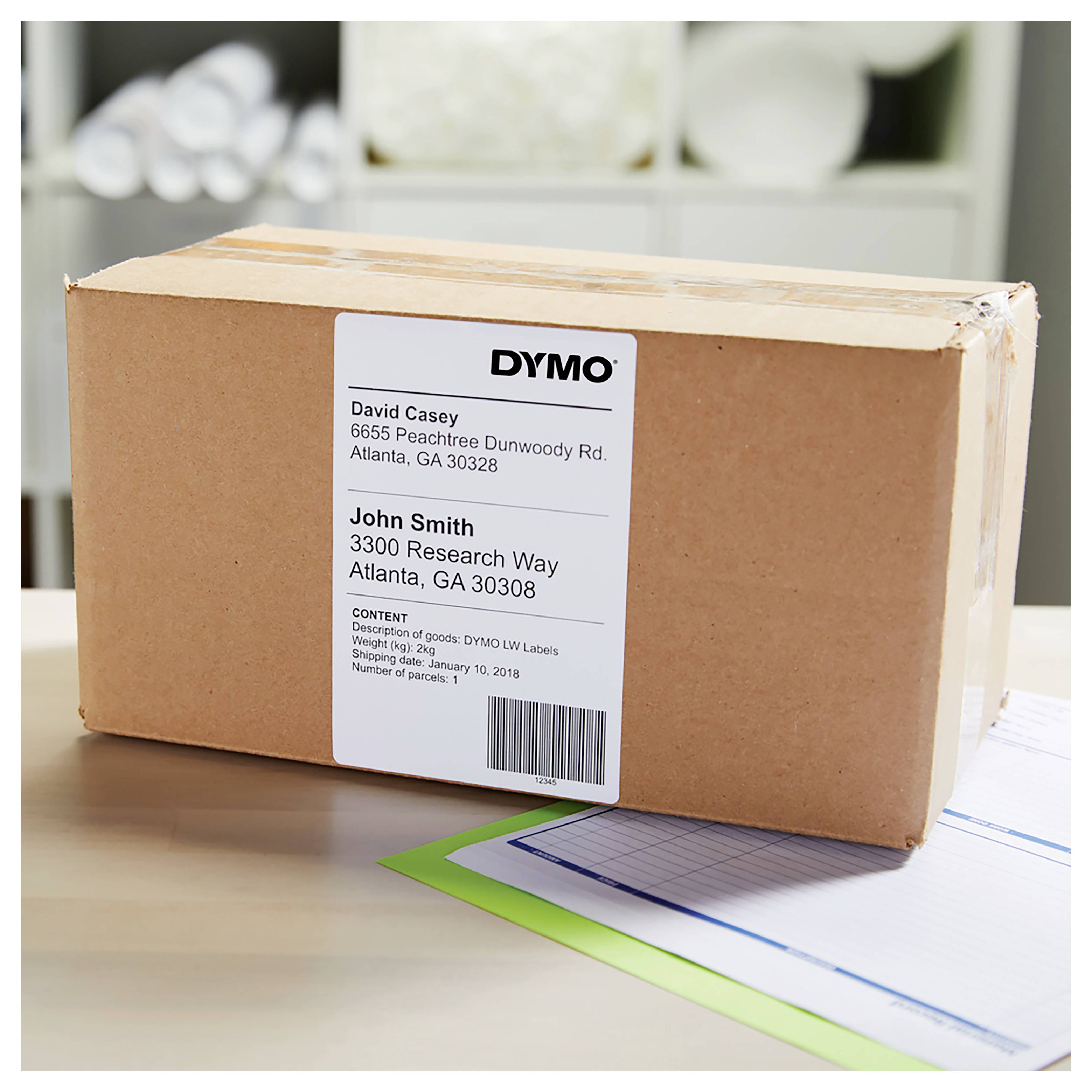 Une boîte d'expédition en carton avec une étiquette d'adresse DYMO pour John Smith sur un bureau, avec des papiers en dessous.