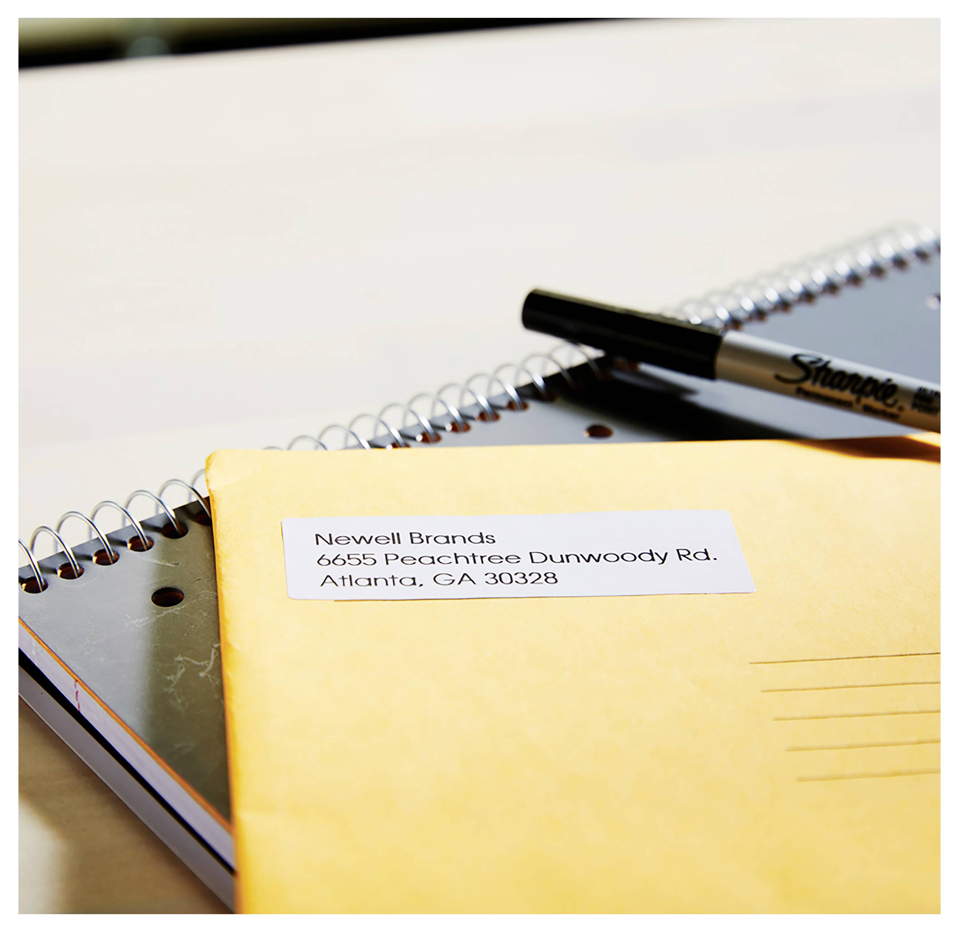 Enveloppe jaune sur un carnet à spirale avec une étiquette postale adressée à « Newell Brands, 6655 Peachtree Dunwoody Rd., Atlanta, GA 30328 » et un marqueur.