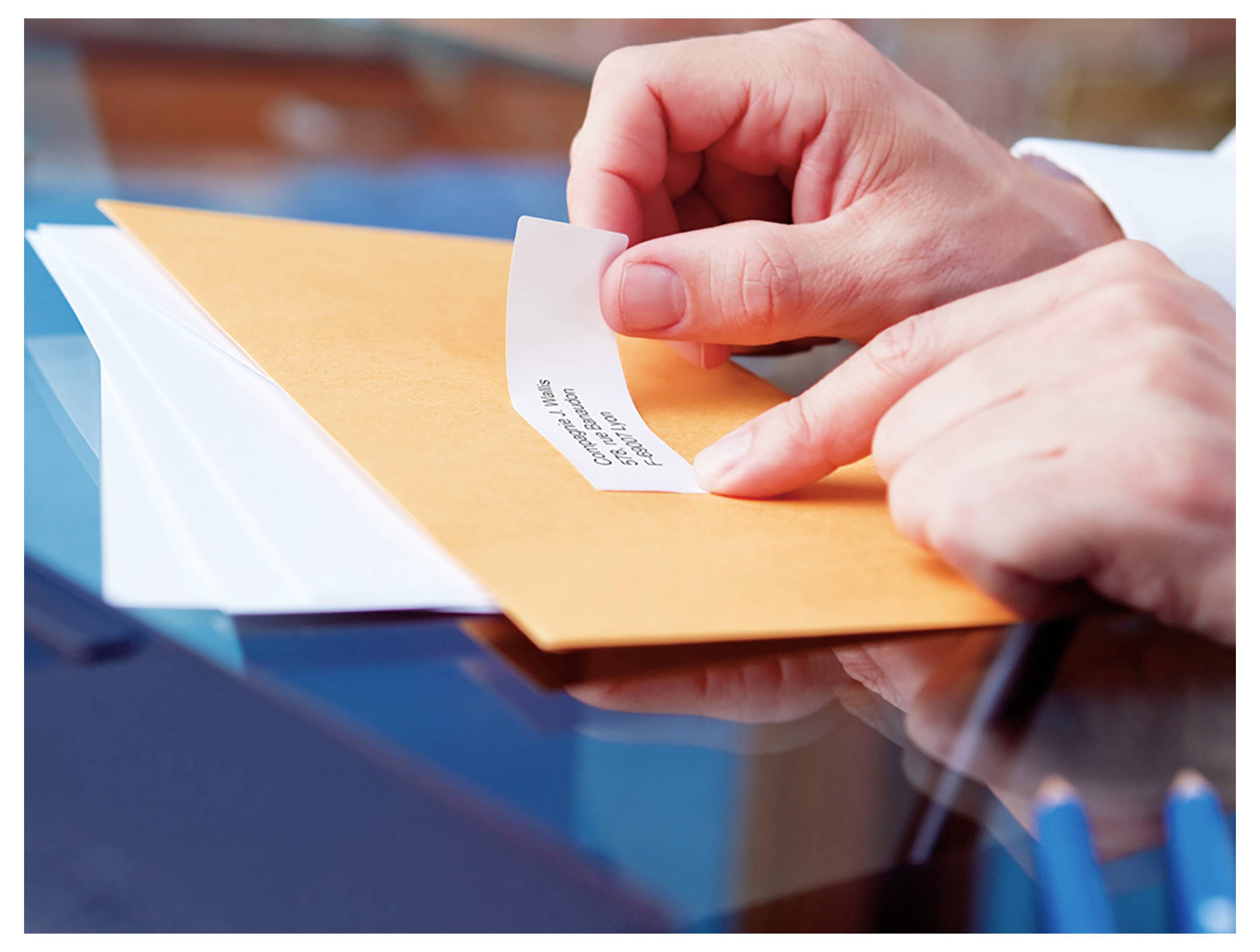 Des mains plaçant un petit morceau de papier dans une enveloppe orange sur une table en verre, suggérant l'envoi ou la fermeture d'un message.