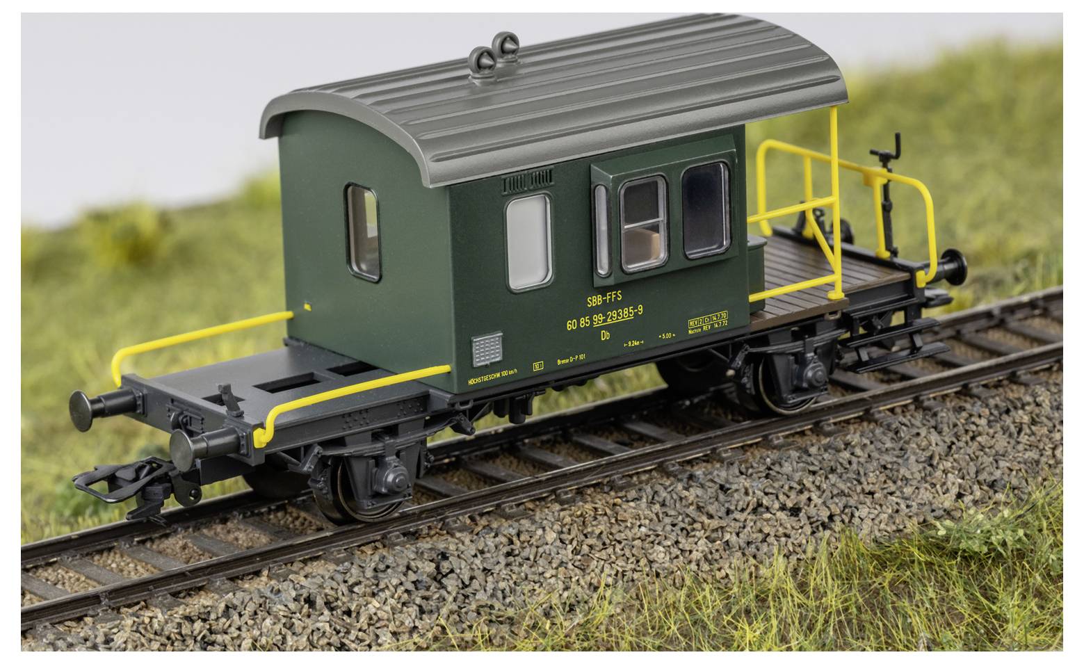 Une cabine de queue de train miniature vert foncé avec un toit gris et des garde-corps jaunes est positionnée sur des rails de chemin de fer à côté de l'herbe.
