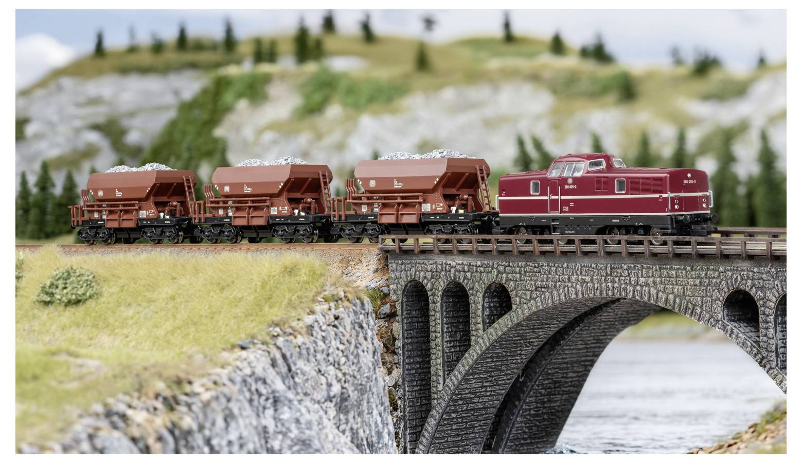 Train miniature avec locomotive rouge et wagons de marchandises traversant un pont en arc de pierre au-dessus de l'eau dans un paysage en miniature.