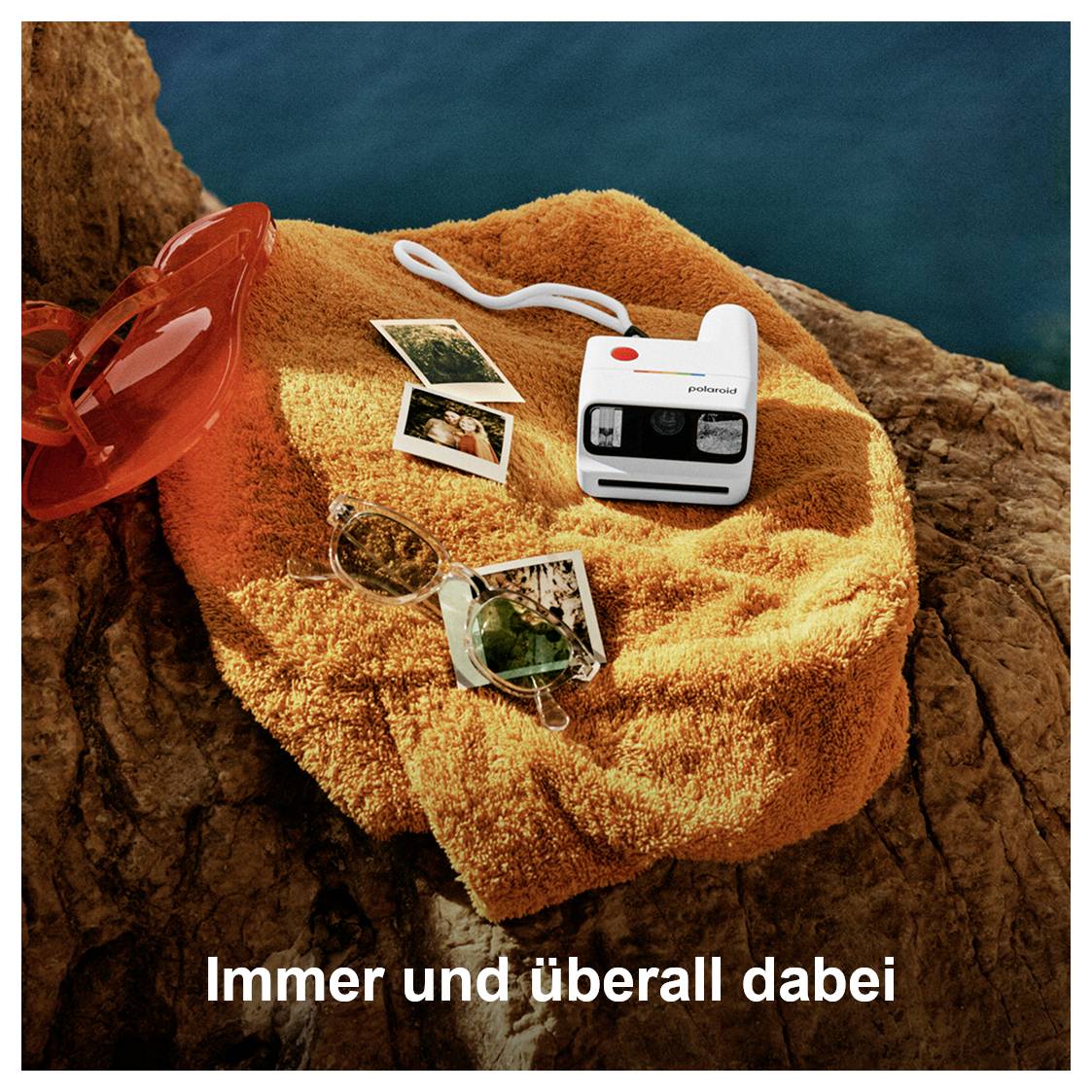 Serviette orange sur des rochers avec un appareil photo Polaroid, des photos instantanées et des lunettes de soleil ; en bas le slogan « Toujours et partout avec toi ».