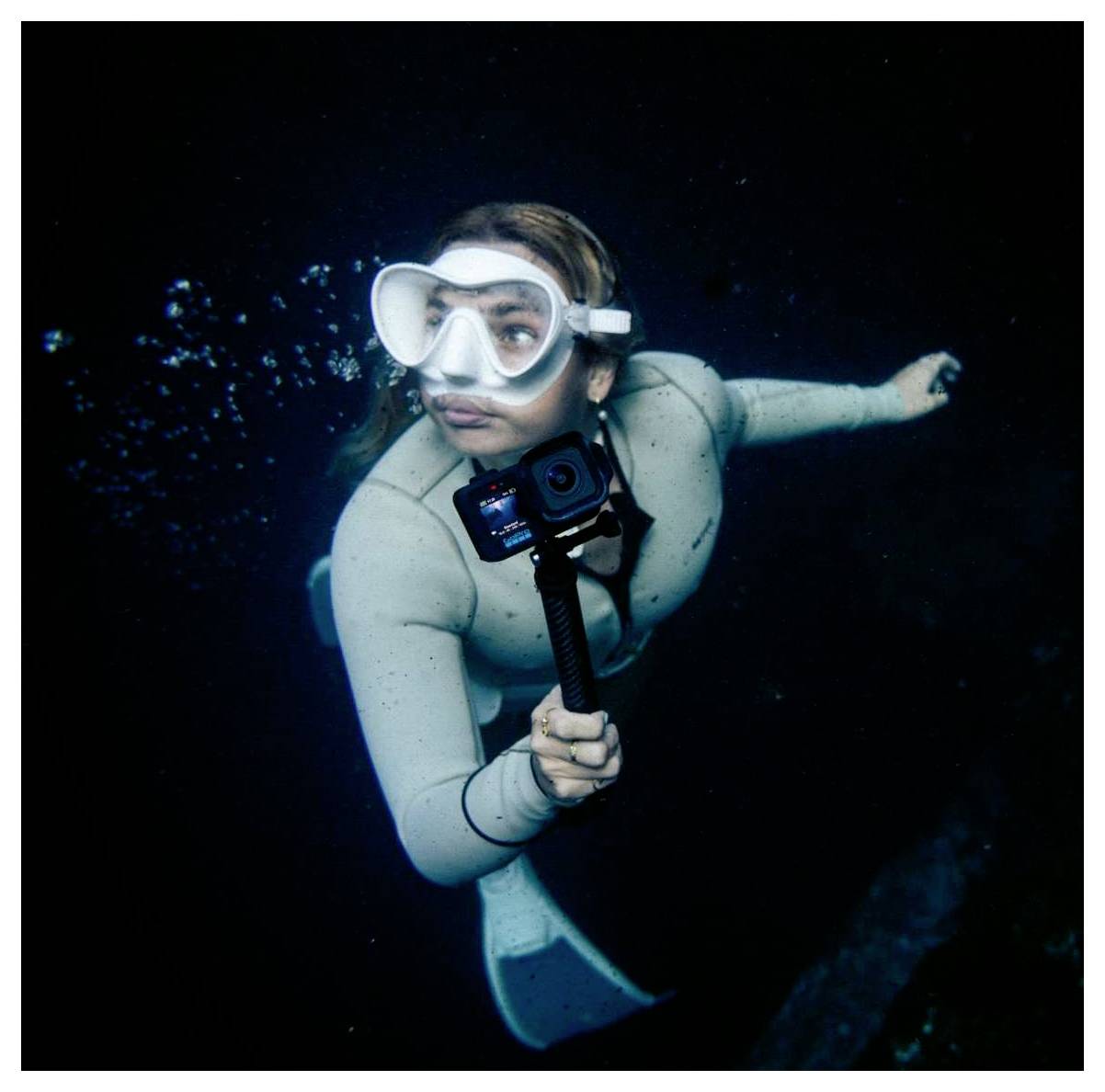 Plongeuse sous-marine en combinaison de plongée et masque tenant un appareil photo sur un bâton à selfie, nageant dans une eau sombre avec des bulles derrière elle.