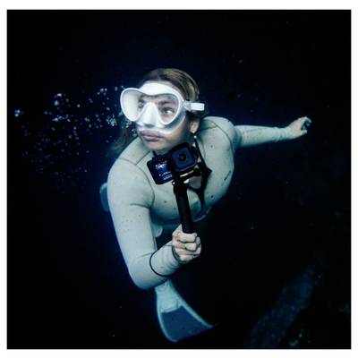 Plongeuse sous-marine en combinaison de plongée et masque tenant un appareil photo sur un bâton à selfie, nageant dans une eau sombre avec des bulles derrière elle.