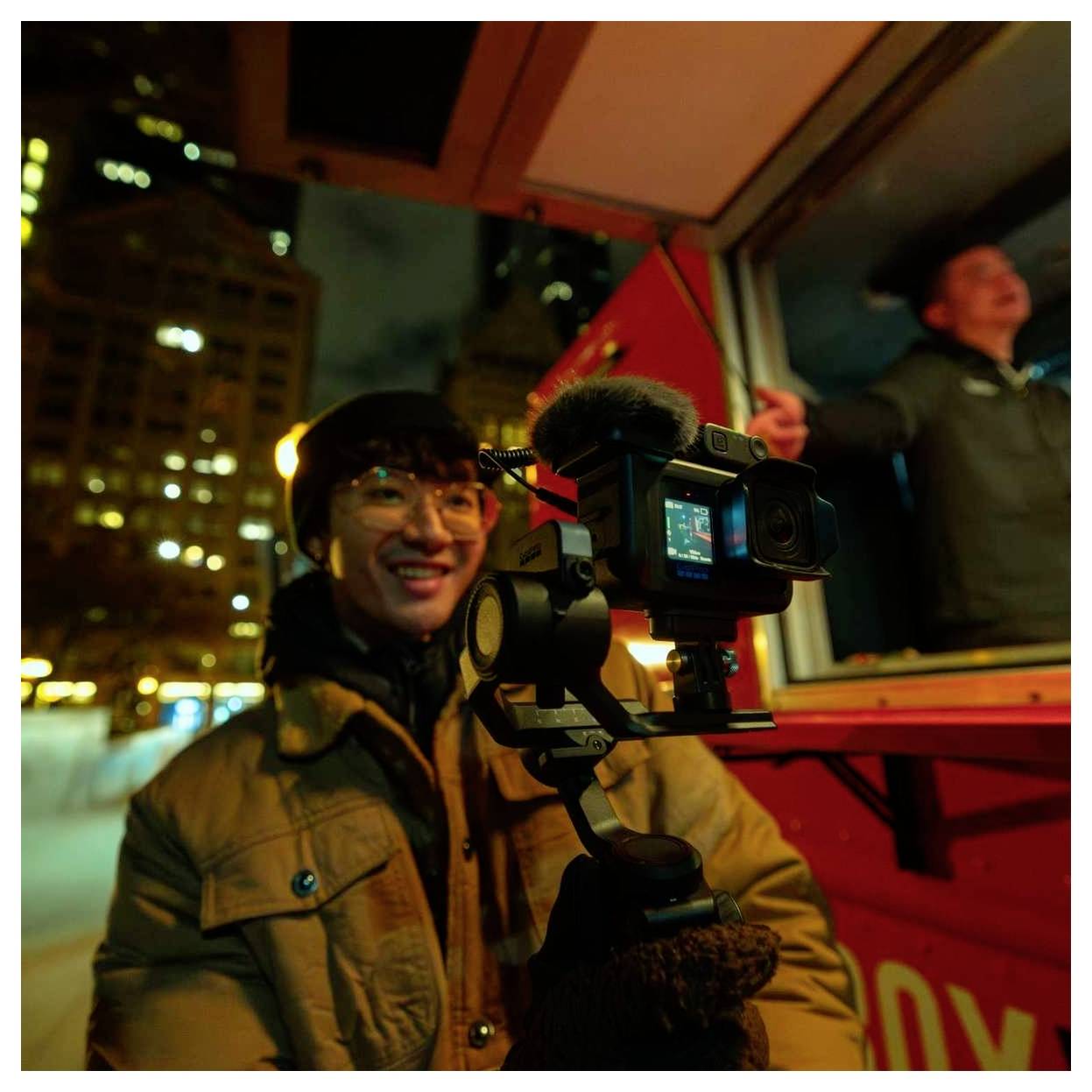 Personne filmant la nuit avec un équipement de caméra sur un véhicule rouge en mouvement, souriante tandis que les lumières de la ville s'estompent en arrière-plan.