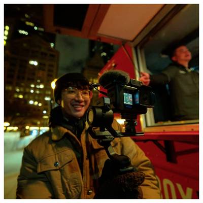 Personne filmant la nuit avec un équipement de caméra sur un véhicule rouge en mouvement, souriante tandis que les lumières de la ville s'estompent en arrière-plan.