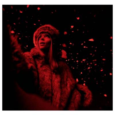 Femme en manteau de fourrure et chapeau se tient dans une lumière rouge contre un fond sombre avec des particules flottantes brillantes.