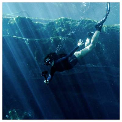 Un tuba sous l'eau près d'un récif rocheux, tenant une caméra et nageant dans une eau bleue illuminée par le soleil.