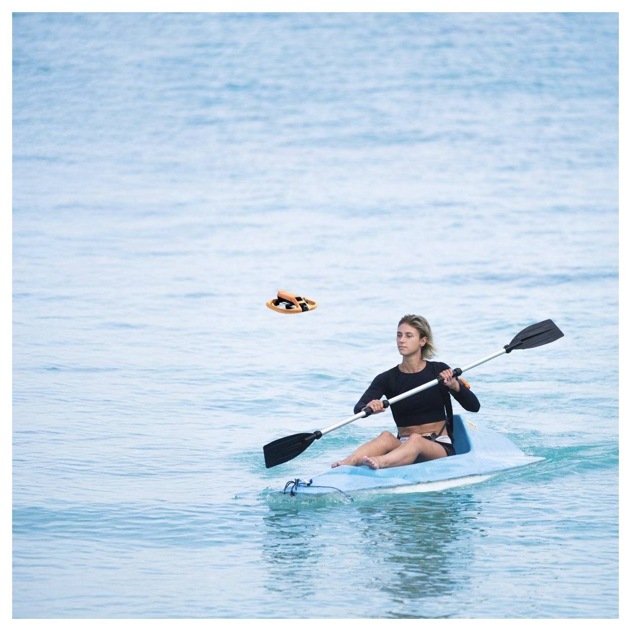 Femme en kayak sur une eau bleue calme, pagayant un kayak bleu clair près d'une petite bouée de sauvetage orange flottante.