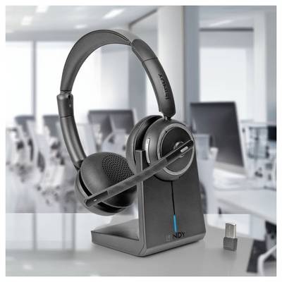 Casque d'écoute noir serre-tête avec microphone à perche sur un socle de recharge, présenté sur un bureau dans un cadre de bureau florisé avec une clé USB à proximité.
