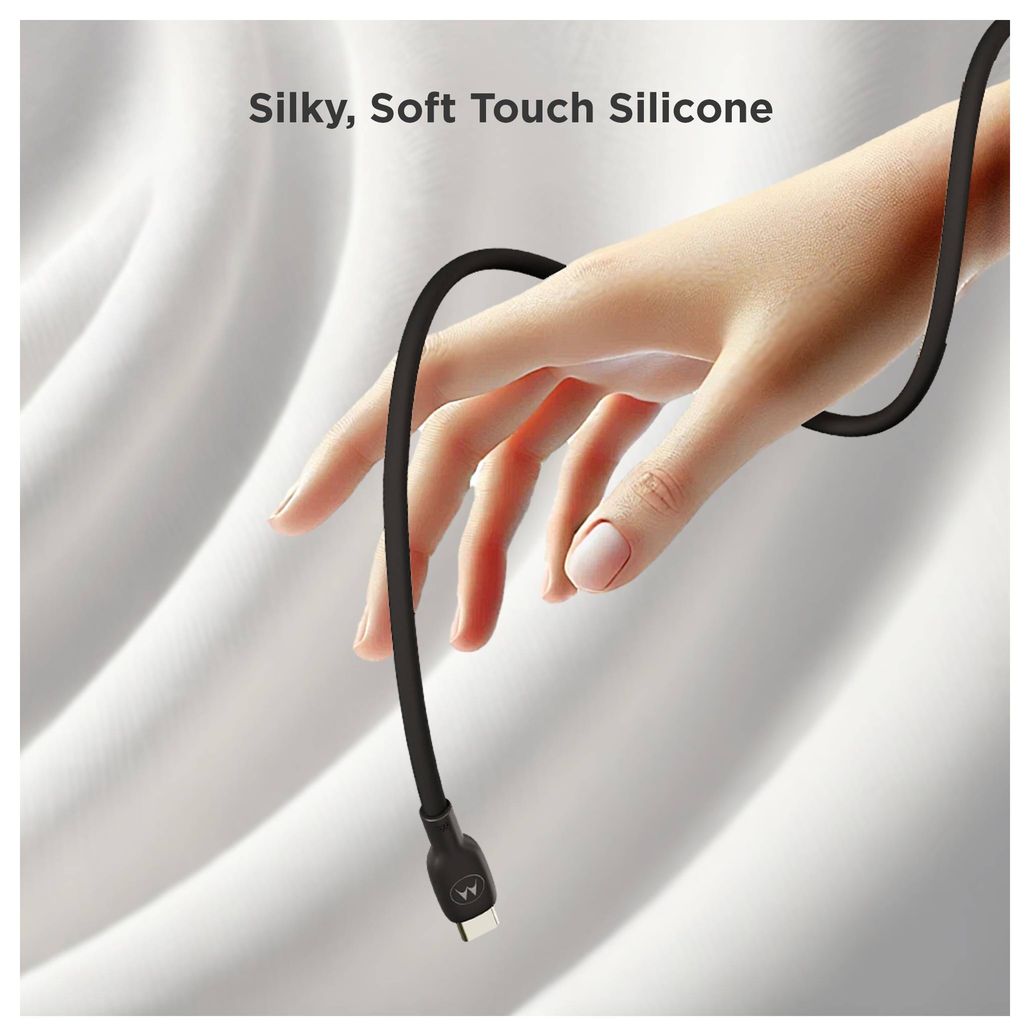 Main tenant un câble de recharge en silicone noir avec le texte « Silky, Soft Touch Silicone » sur un fond de tissu clair.