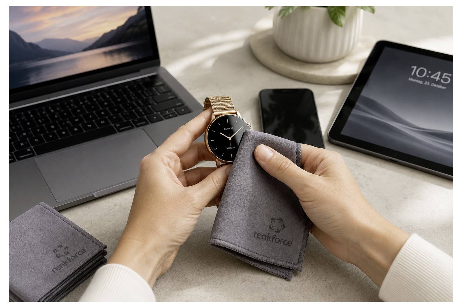 Des mains tenant une montre intelligente et un chiffon de nettoyage à côté d'un ordinateur portable, d'un téléphone et d'une tablette sur un bureau, suggérant le nettoyage d'écran ou d'appareil.